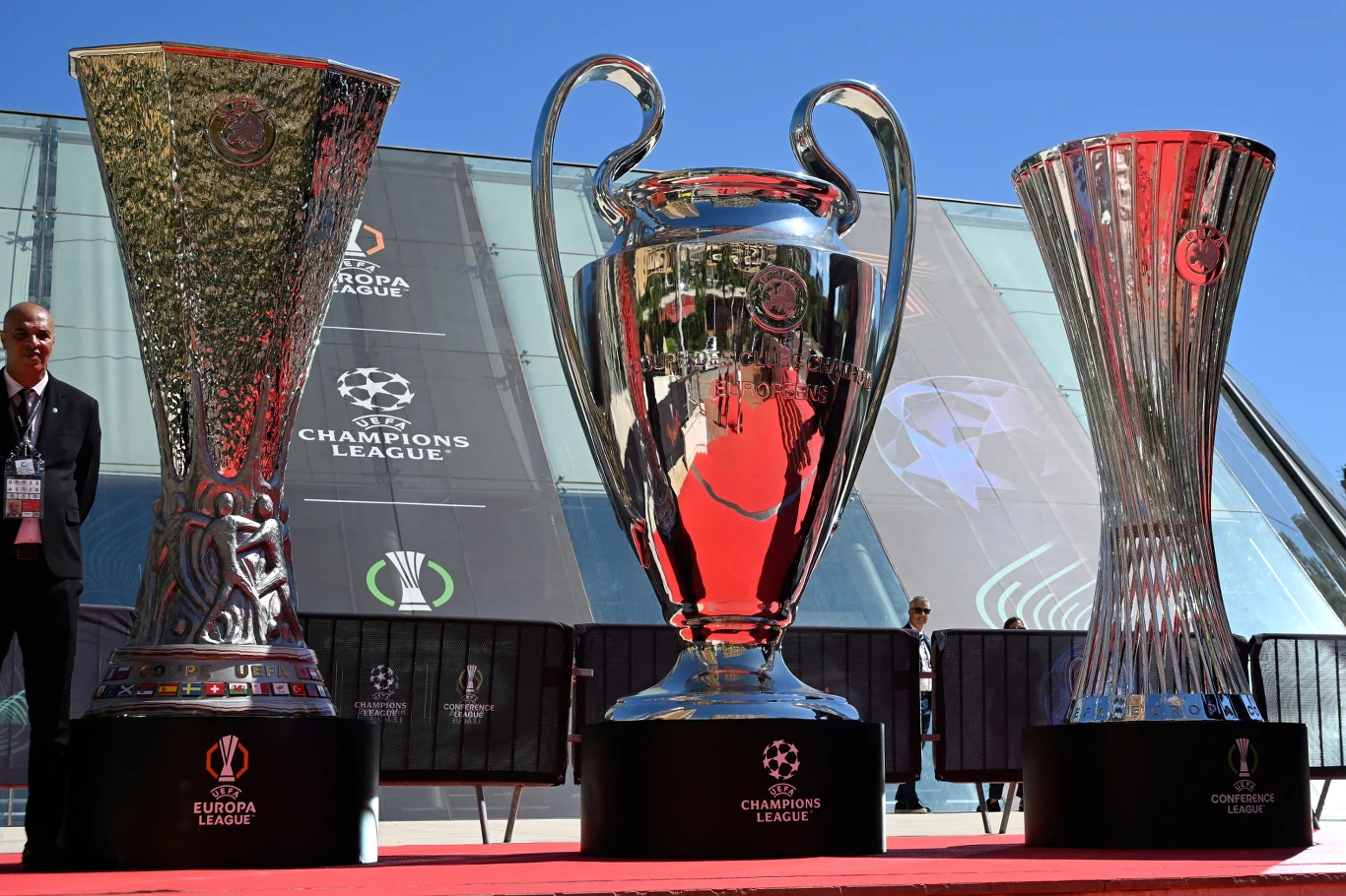 Puchary Ligi Europy, Ligi Mistrzów i Ligi Konferencji Trzy prestiżowe trofea piłkarskie ustawione na czerwonym dywanie, w centrum puchar Ligi Mistrzów UEFA, po bokach puchar Ligi Europy oraz puchar Ligi Konferencji, w tle przeszklona fasada budynku i logo Champions League.