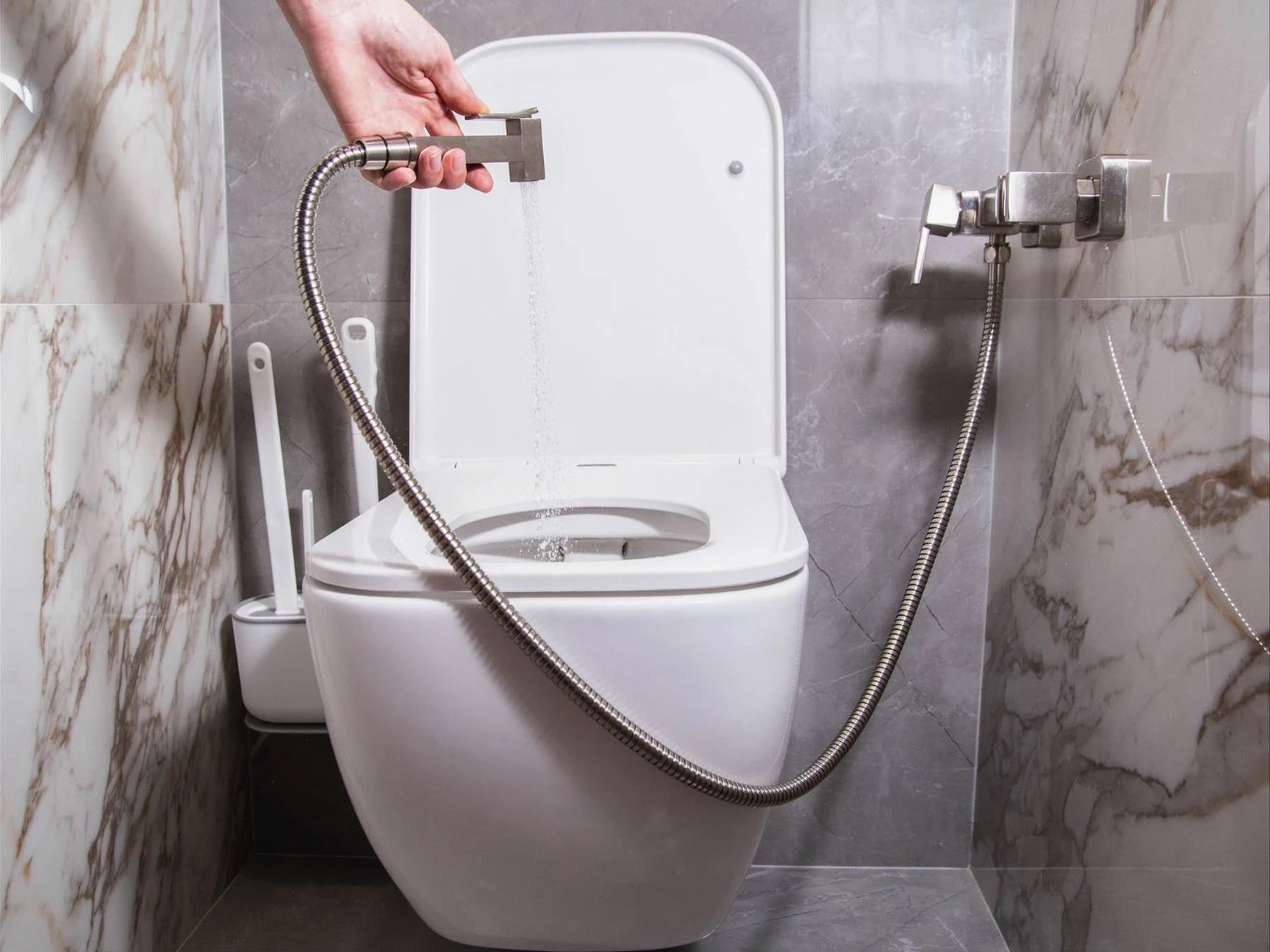 Toaleta z otwartą klapą, w której wnętrzu skierowana jest ręczna słuchawka bidetowa trzymana przez osobę, obok widoczne akcesoria łazienkowe, kafelki na ścianach w odcieniach szarości i bieli.