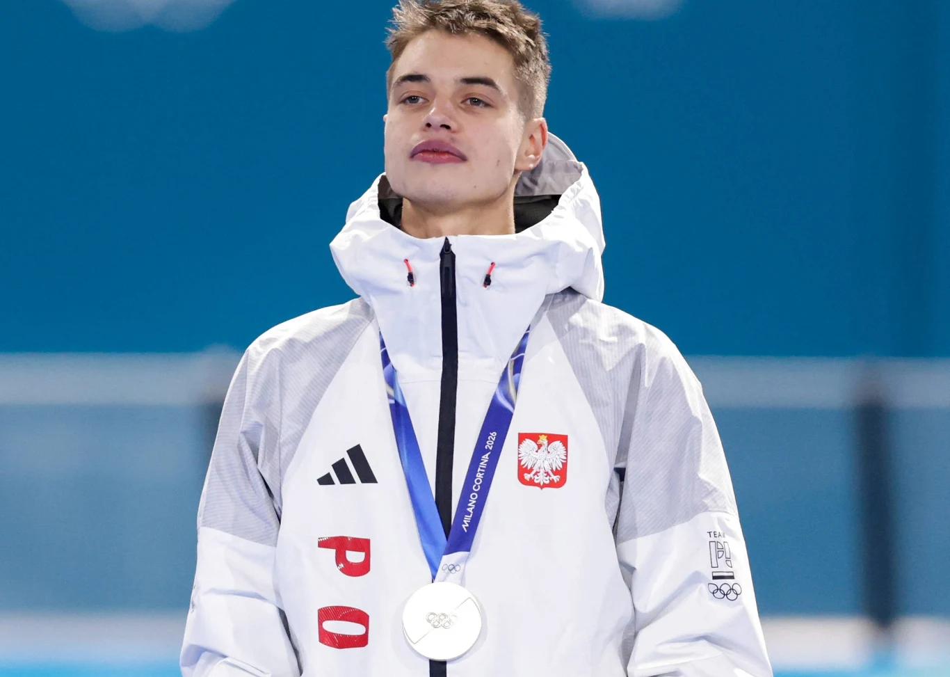 Władimir Semirunnij Sportowiec w białej kurtce z emblematem Polski stoi na podium z zawieszonym na szyi srebrnym medalem olimpijskim, na niebieskim tle.