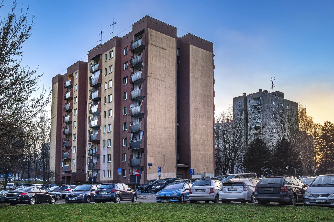 Obowiązek zapłaty podatku gruntowego dotyczy właścicieli mieszkań, którzy posiadają udział w nieruchomości wspólnej Wielokondygnacyjny blok mieszkalny z balkonami otoczony zaparkowanymi samochodami, w tle drugi budynek, drzewa oraz niebieskie niebo