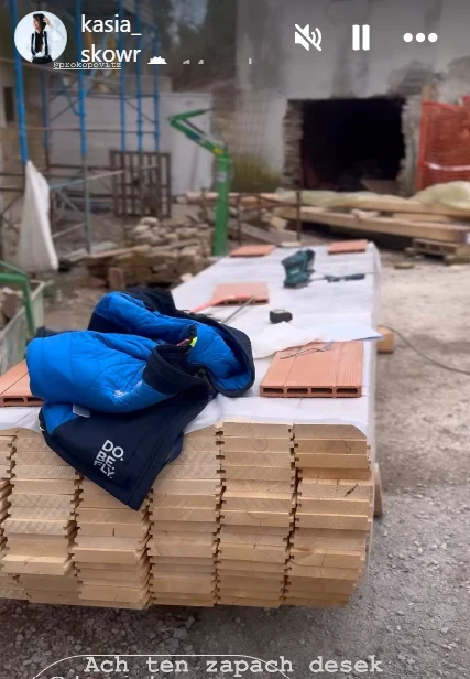 Stos desek drewnianych ułożonych na placu budowy, na których leży niebieska kurtka i narzędzia, w tle widoczne elementy konstrukcyjne oraz niezakończony budynek.