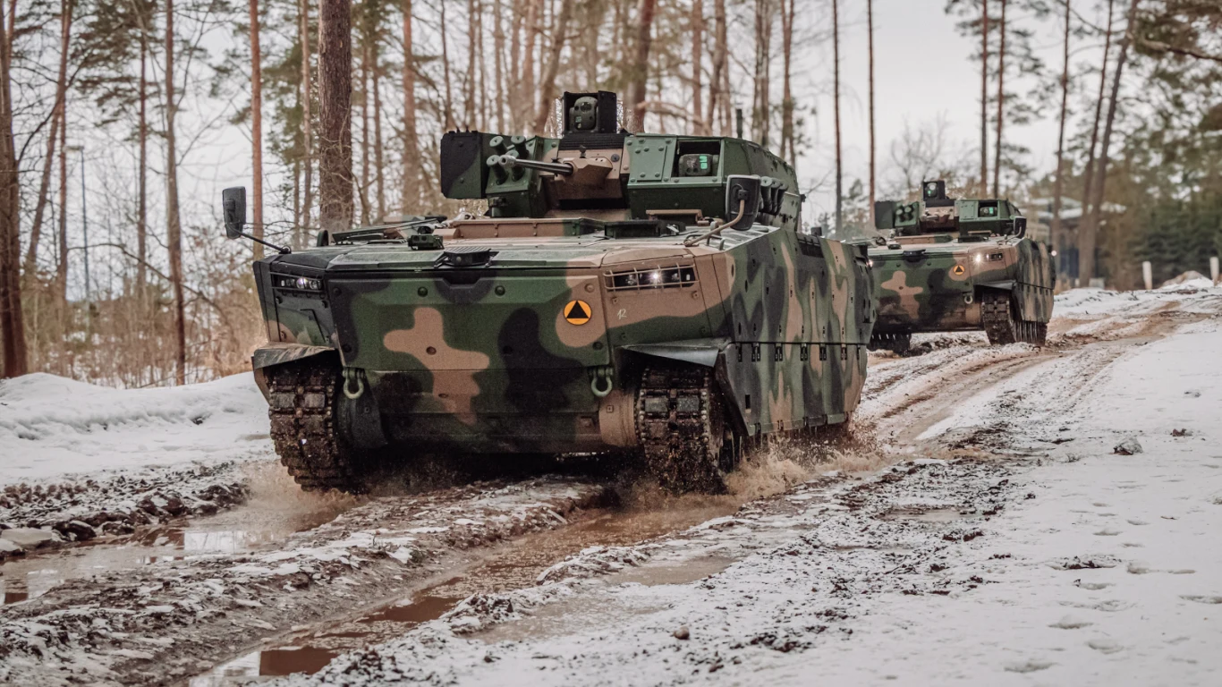 Dwa opancerzone pojazdy wojskowe z kamuflażem leśnym jadą leśną, błotnistą drogą w zimowej scenerii, otoczone wysokimi drzewami.