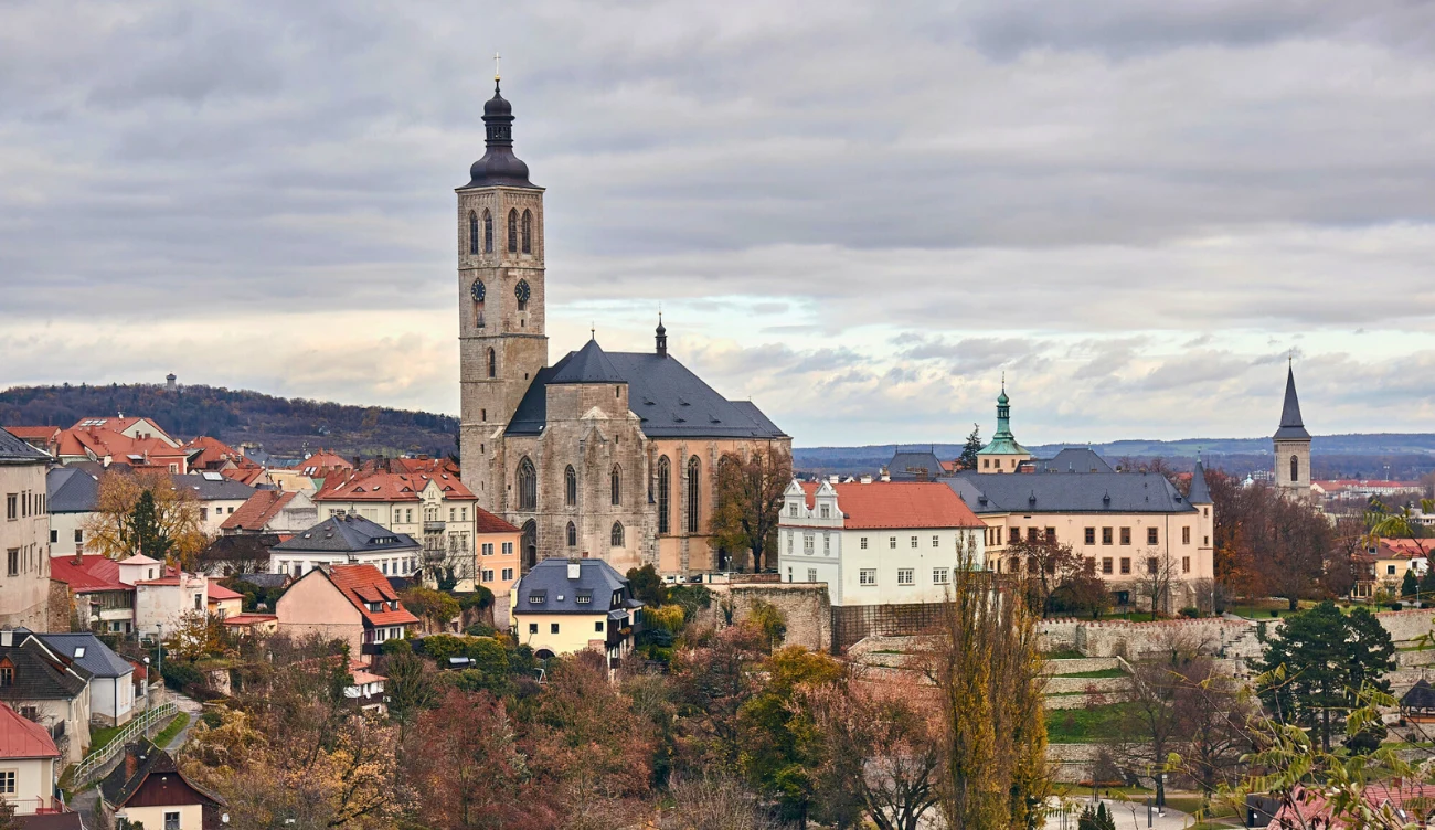 Gdzie jest Kutna Hora? Dominuje panorama miasta z wyróżniającym się dużym, historycznym kościołem o wieży górującej nad okolicznymi budynkami, w otoczeniu jesiennych drzew i rozległego krajobrazu z niskimi domami.