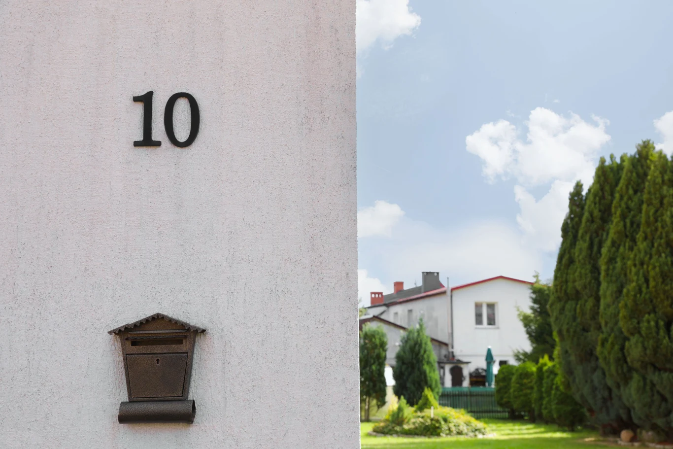 Biała ściana budynku z numerem 10 i skrzynką pocztową na pierwszym planie, w tle ogród oraz dom otoczony zielonymi drzewami pod niebieskim niebem z kilkoma chmurami.