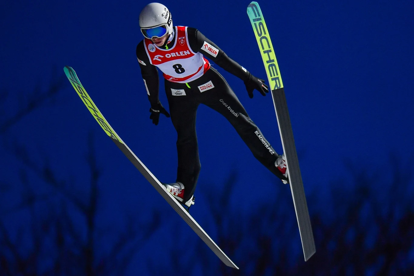Konrad Tomasiak Skoczek narciarski w powietrzu, ubrany w czarny kombinezon z numerem startowym 8, kask ochronny i gogle, wysokie narty z napisem Fischer na tle ciemnoniebieskiego nieba.