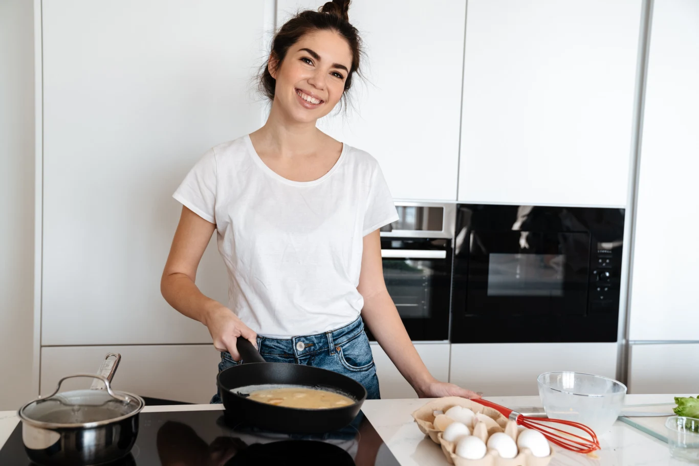 Dla osób spod znaku Raka jedzenie stanowi wyraz opieki Młoda kobieta w białej koszulce i dżinsach smaży jajka na patelni w nowoczesnej kuchni, obok niej leżą jajka, trzepaczka i miseczka.