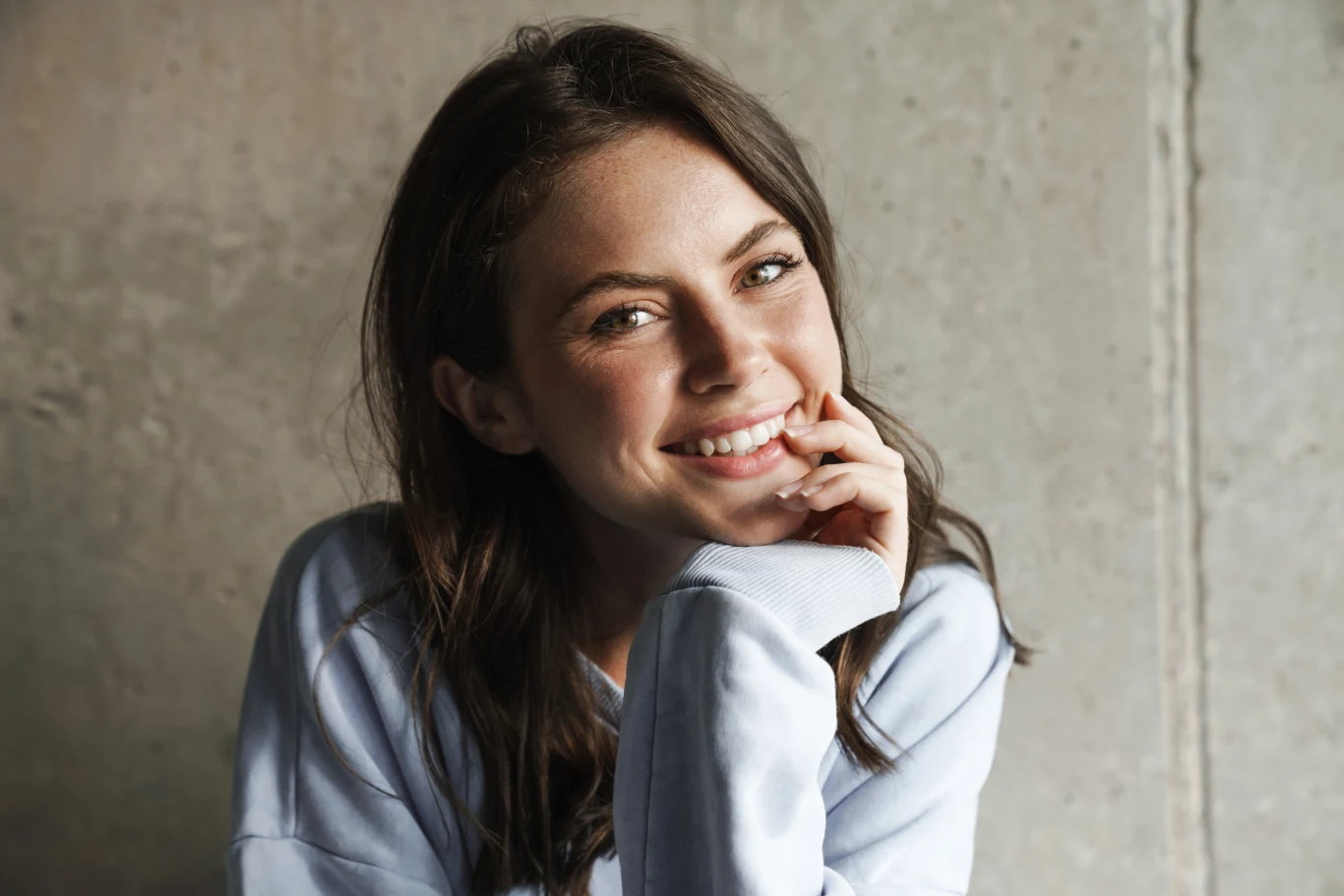 Młoda kobieta z ciemnymi włosami uśmiecha się, opierając głowę na dłoni, ubrana w jasną bluzę, siedzi na tle szarej ściany.