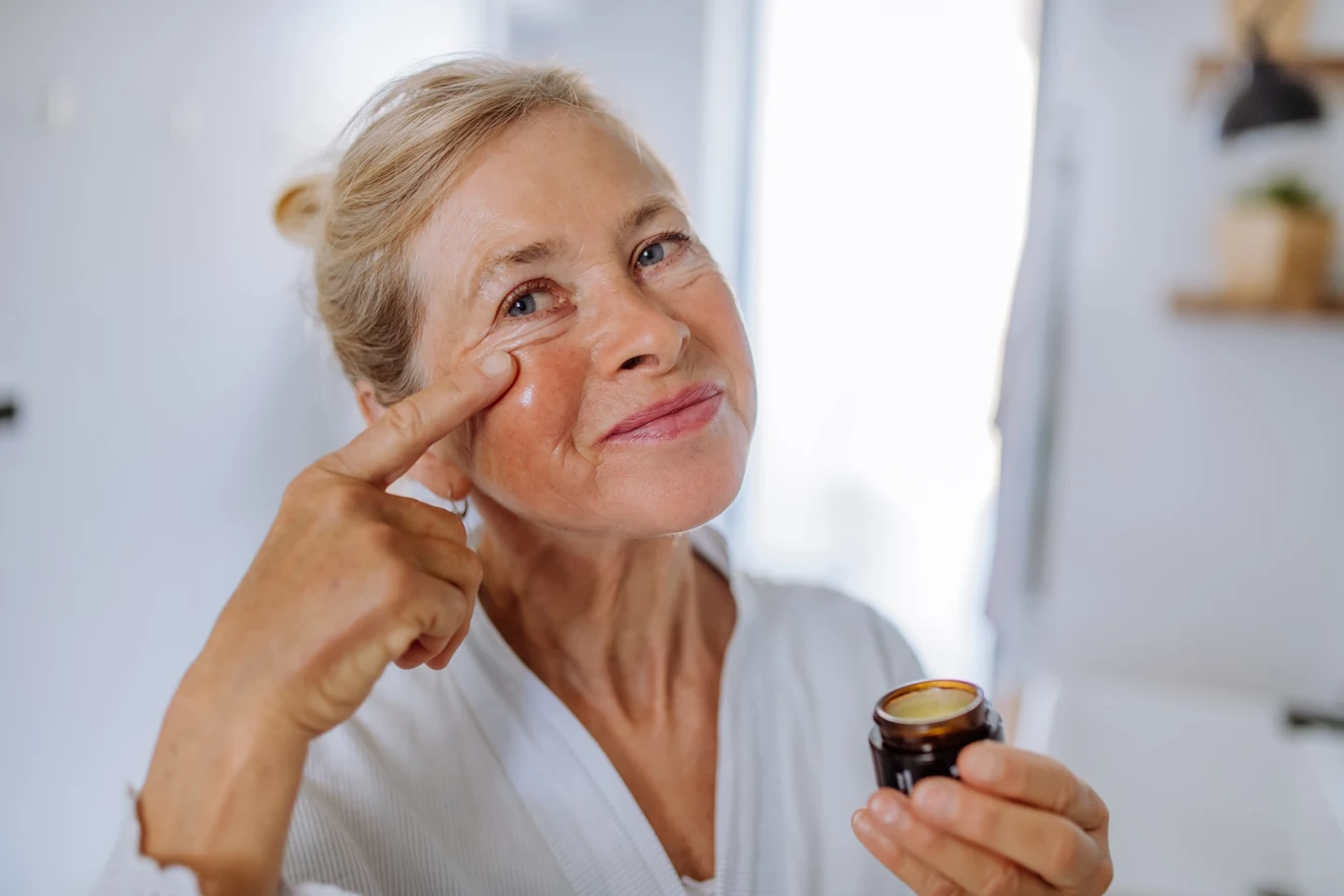 Działąją silniej niż krem anti-aging. Kup w aptece, stosuj 3 razy w tygodniu Starsza kobieta w jasnym szlafroku nakłada krem na skórę pod oczami, uśmiechając się delikatnie do obiektywu.