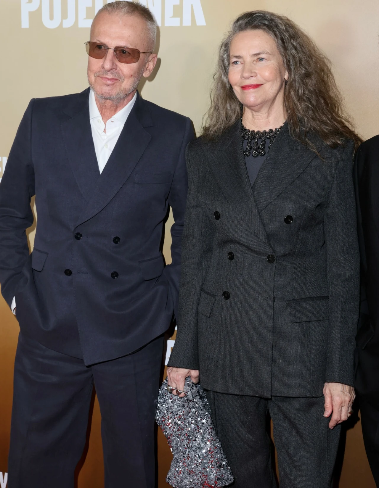 Elegancka para w średnim wieku ubrana w stylowe garnitury, kobieta z długimi włosami trzyma srebrną torebkę, oboje zadowoleni, stoją na tle złotej ścianki z napisem.