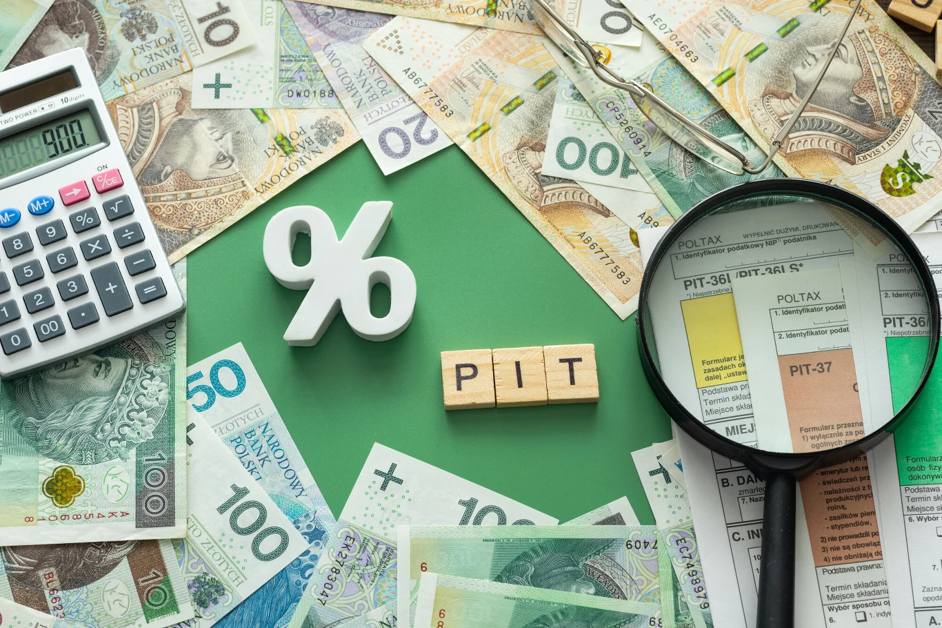 Kiedy otrzymam zwrot podatku? Poznaj kluczowe terminy Banknoty polskie, kalkulator, symbol procentu, drewniane kostki z napisem PIT oraz formularze podatkowe i lupa tworzą kompozycję nawiązującą do tematyki podatków dochodowych i rozliczeń finansowych.