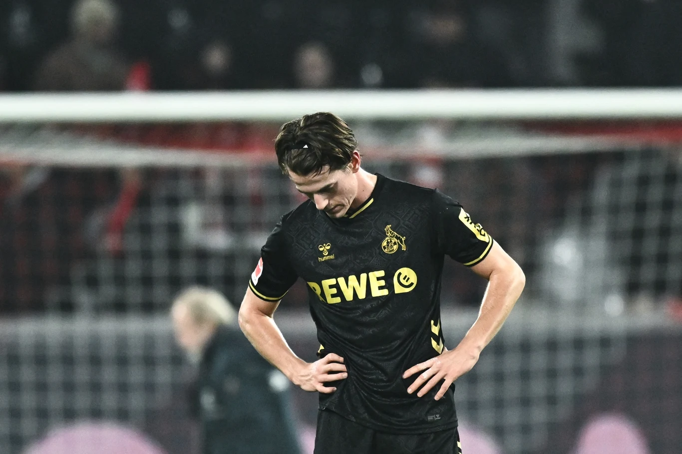 Jakub Kamiński Piłkarz w czarnym stroju z emblematem klubu piłkarskiego FC Köln stoi ze spuszczoną głową i rękami opartymi na biodrach na tle nieostrego stadionu, wyrażając rozczarowanie lub smutek.