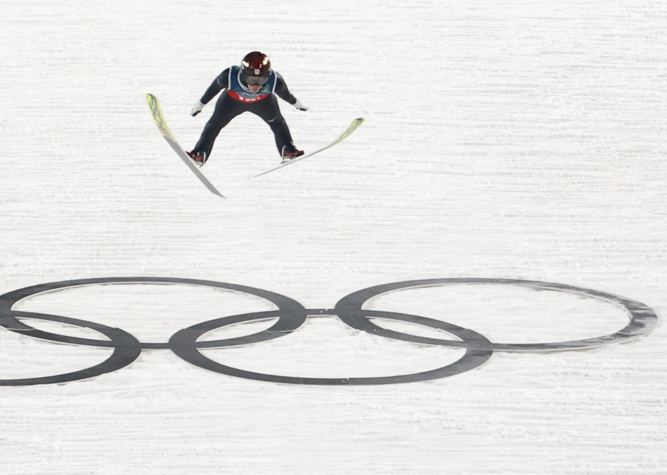 Skoki narciarskie. Ren Nikaido w locie Skoczek narciarski podczas lotu nad śniegiem, poniżej widoczne są olimpijskie koła na białej powierzchni skoczni.