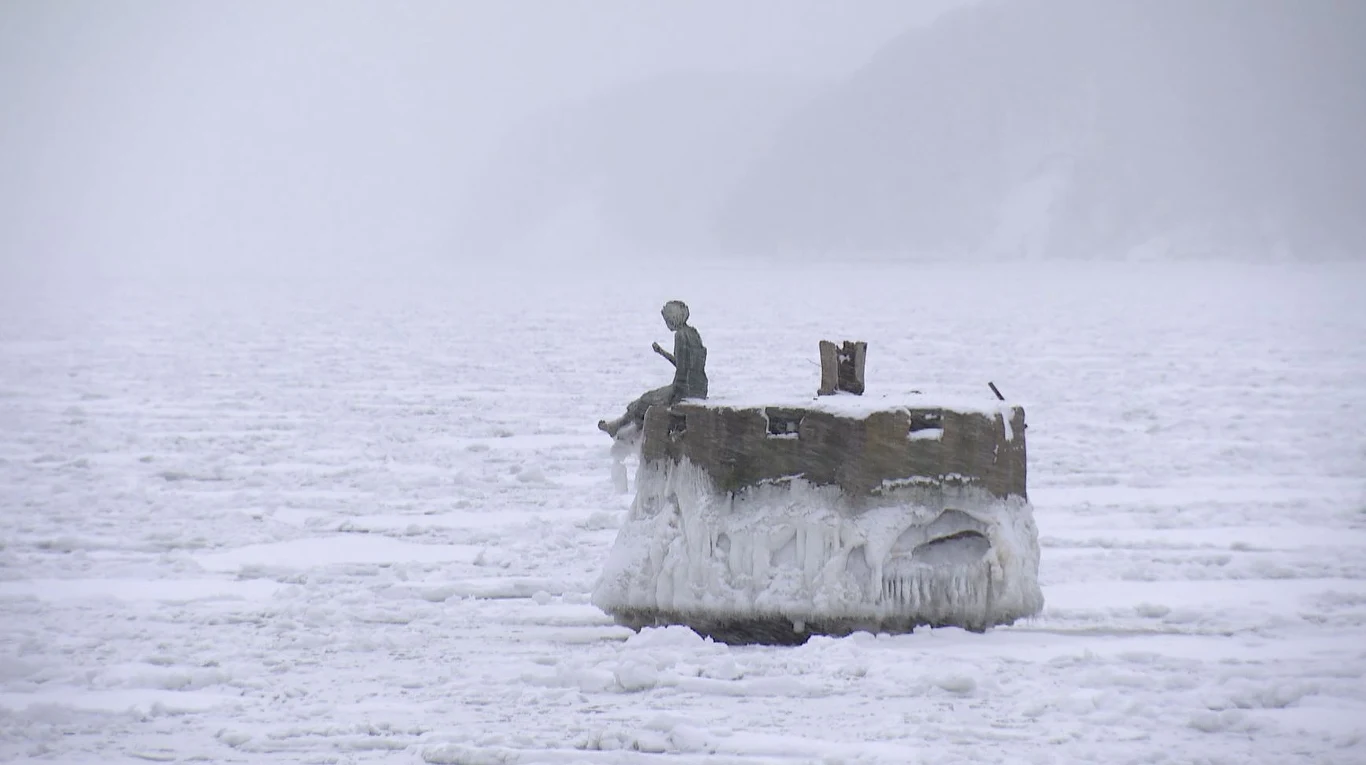 Zamarznięta rzeźba siedzącej postaci na betonowym bloku pokrytym grubą warstwą lodu, otoczona śniegiem i lodem na rozległej, zimowej przestrzeni.