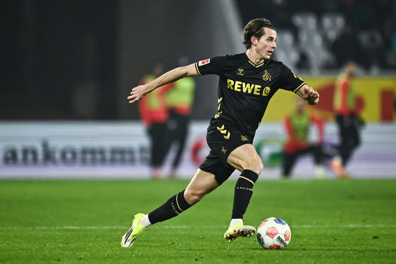 Jakub Kamiński Piłkarz w czarnym stroju z żółtymi detalami biegnie z piłką po murawie stadionu podczas meczu piłkarskiego. W tle widoczni są sędziowie oraz rozmyte trybuny.