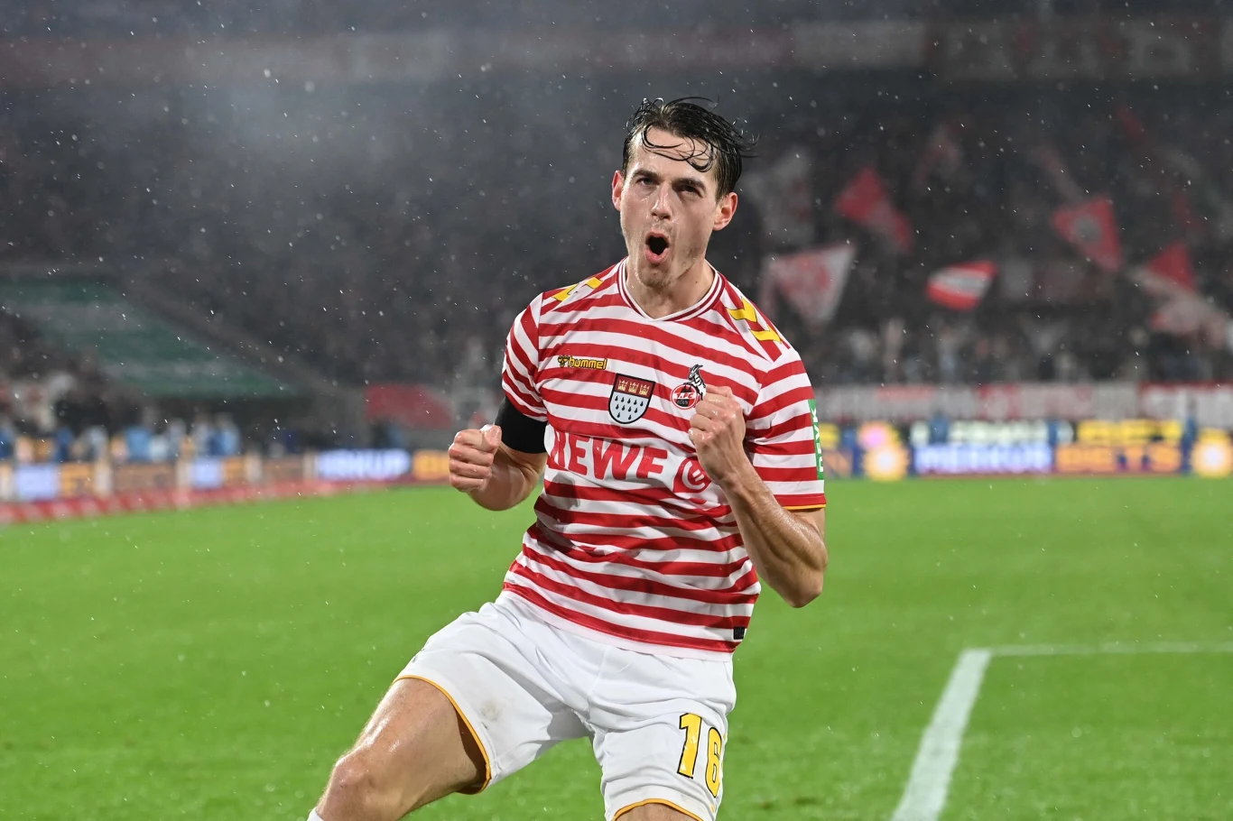 Jakub Kamiński Piłkarz w pasiastym czerwono-białym stroju cieszy się z gola na stadionie podczas meczu piłkarskiego w trakcie deszczu, unosząc zaciśniętą pięść w geście triumfu, w tle widoczne trybuny z kibicami.