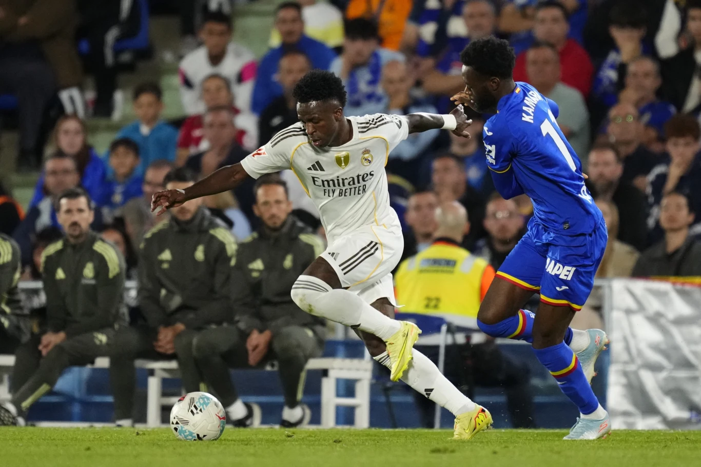 Vinicius Junior podczas meczu La Liga Real Madryt - Getafe Dwóch piłkarzy rywalizuje o piłkę podczas meczu, jeden zawodnik w białym stroju z Realu Madryt biegnie z piłką, drugi piłkarz w niebieskim stroju próbuje go zatrzymać, w tle ławka rezerwowych i kibice.