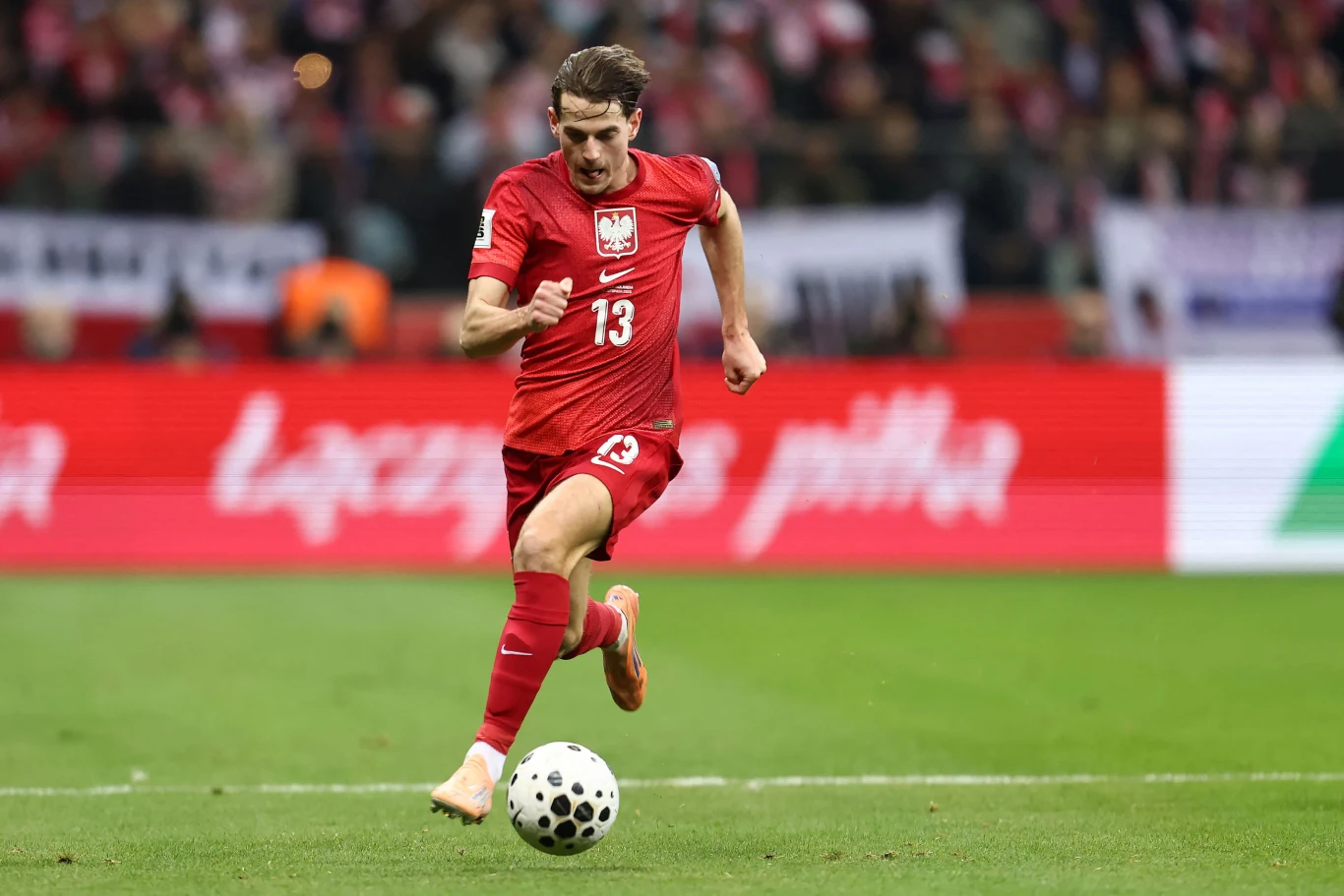 Jakub Kamiński w reprezentacji Polski Polski piłkarz w czerwonym stroju biegnący z piłką po murawie stadionu podczas meczu, w tle widać trybuny pełne kibiców.