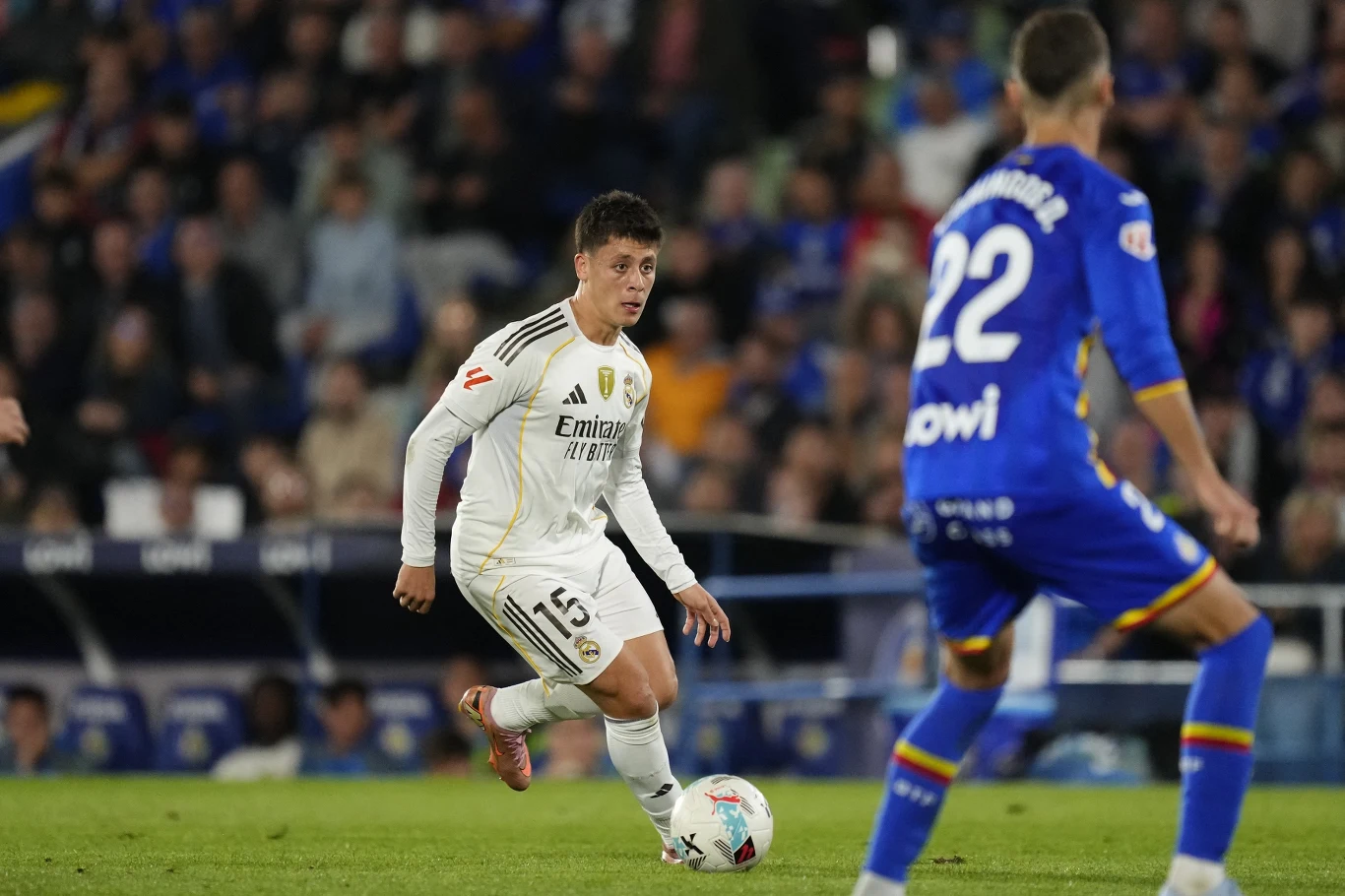 Arda Guler w barwach Realu Madryt podczas meczu La Liga z Getafe Piłkarz w białym stroju kontroluje piłkę podczas meczu, drugi zawodnik w niebieskiej koszulce próbuje go zatrzymać, a w tle widoczny jest tłum kibiców na trybunach.