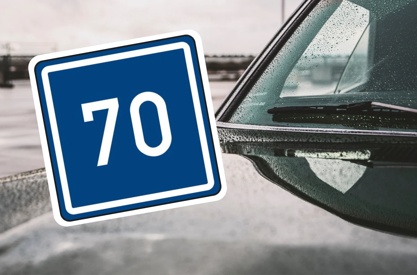 Co oznacza niebieski znak z liczbą? Droga asfaltowa mokra od deszczu, fragment samochodu z widoczną szybą i lusterkiem bocznym, na pierwszym planie znak informacyjny z oznaczeniem ograniczenia prędkości do 70 km/h.