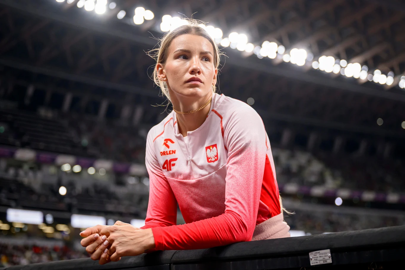 Kobieta ubrana w biało-czerwony dres z orłem i logotypami sponsorów, oparta o barierkę na stadionie lekkoatletycznym, w tle trybuny i oświetlenie stadionowe.