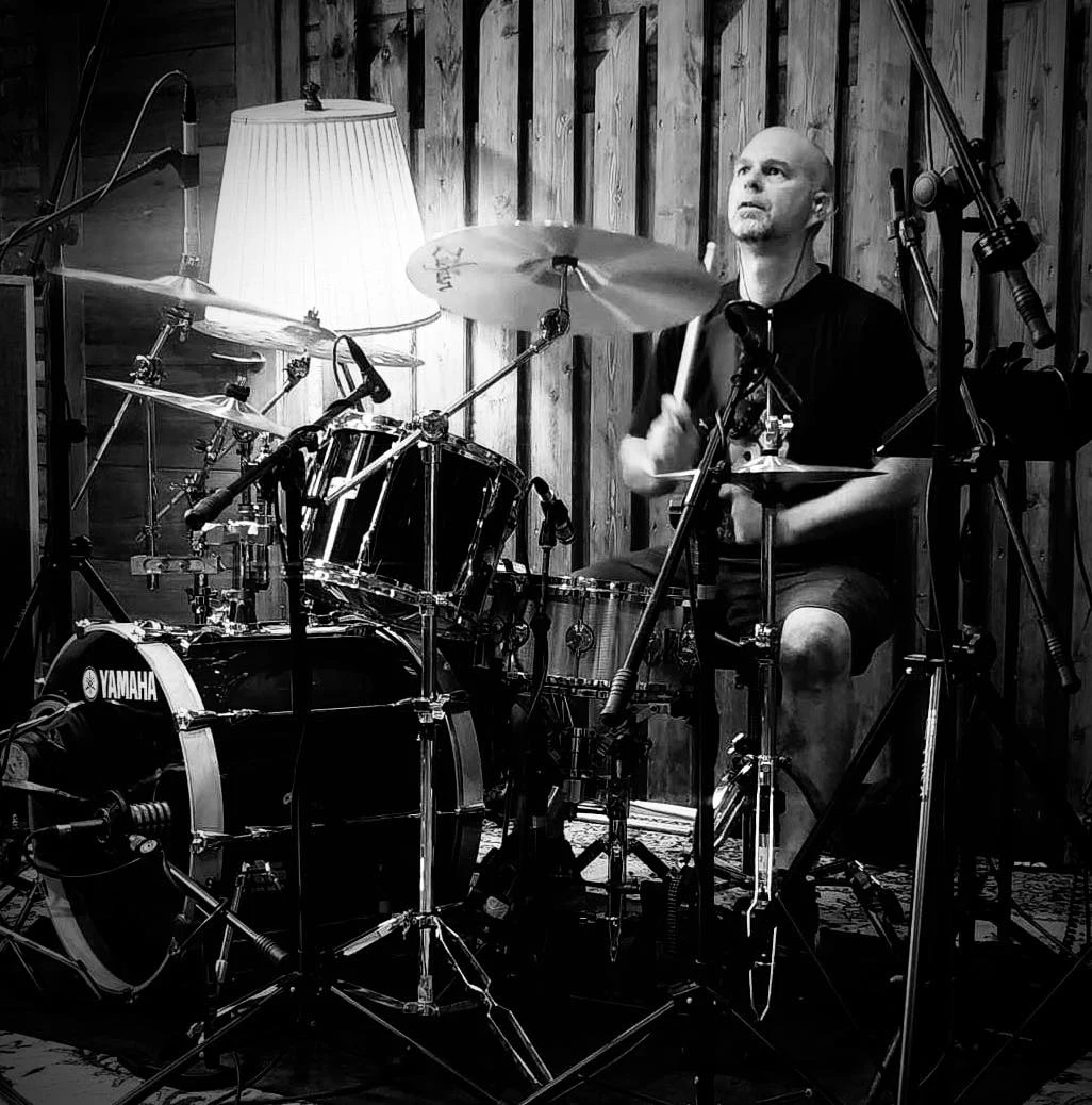 Kamil Filipczak był perkusistą grupy Prawda Mężczyzna grający na perkusji w profesjonalnym studiu nagraniowym, otoczony mikrofonami i wyposażeniem muzycznym, przy lampie i ścianie z drewnianych paneli.