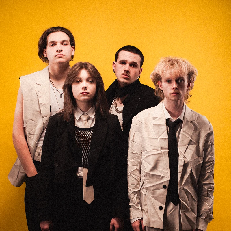 Czworo młodych ludzi ubranych w jasne oraz ciemne, oryginalne stroje, stoi razem na tle żółtej ściany przyjmując poważne wyrazy twarzy.