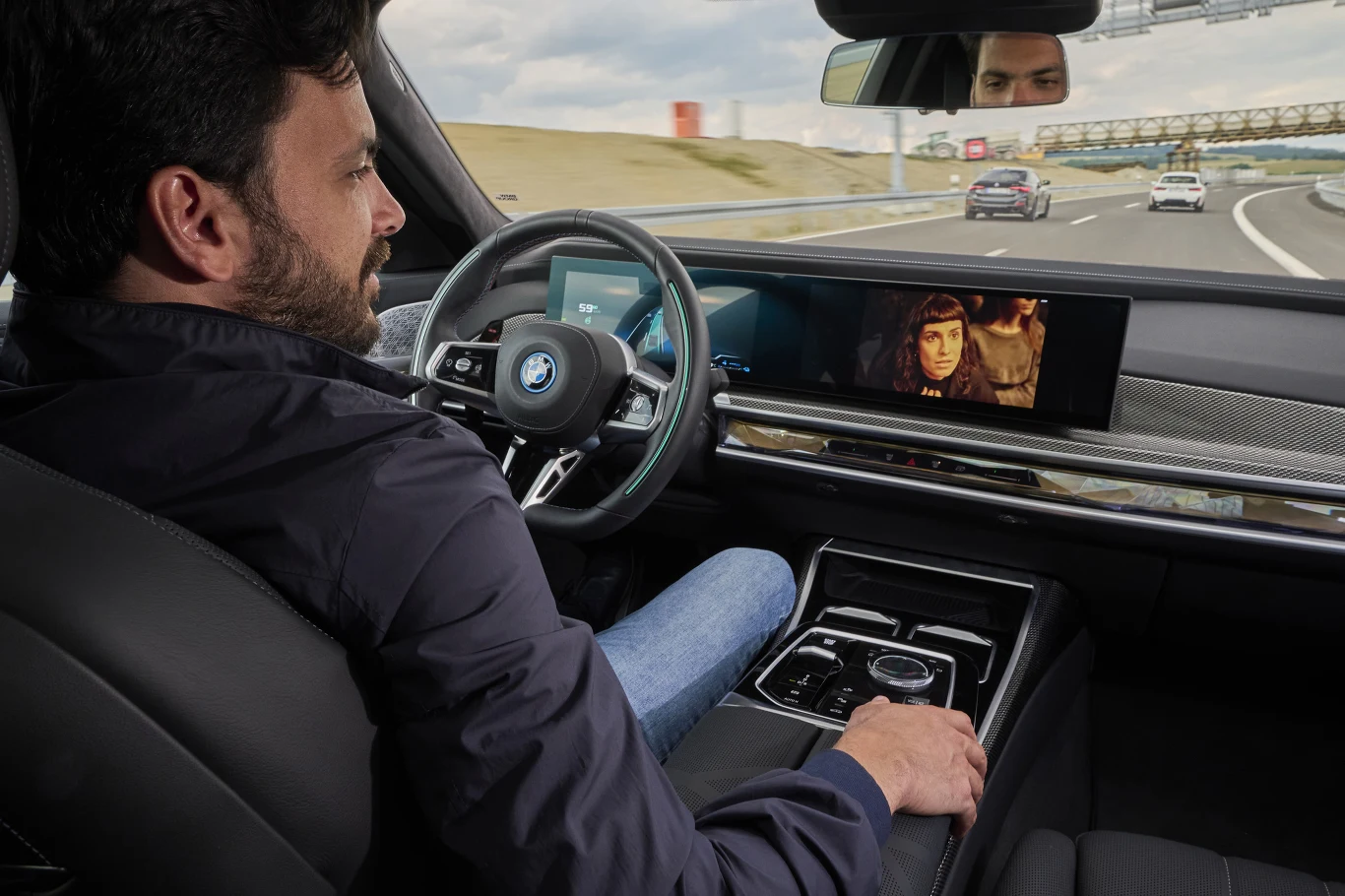 Mężczyzna siedzący za kierownicą samochodu BMW na autostradzie, z ręką opartą na podłokietniku, spogląda na ekran wyświetlający scenę z filmu, podczas gdy pojazd porusza się po drodze obok innych aut.