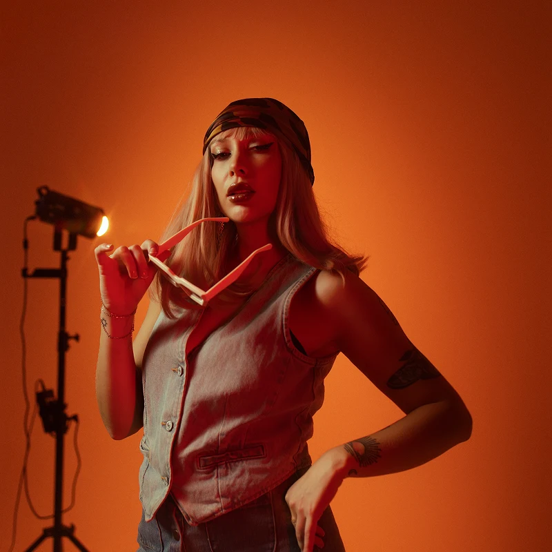 Kobieta w kamizelce jeansowej i bandanie na głowie, trzymająca okulary przeciwsłoneczne, pozuje na tle pomarańczowego tła z profesjonalnym oświetleniem studyjnym.