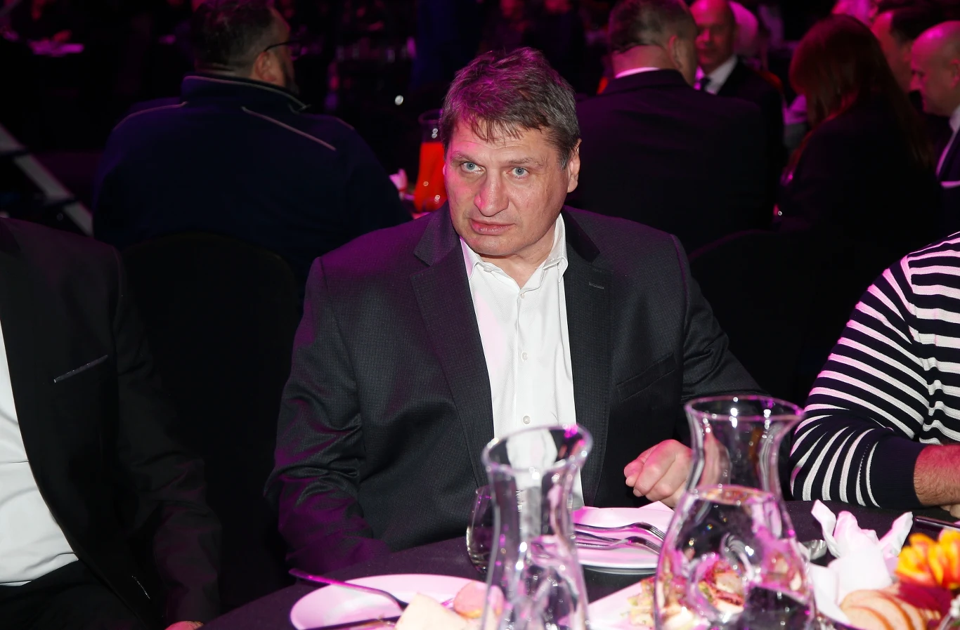 Mężczyzna w garniturze siedzący przy stole na wydarzeniu, wokół niego inni uczestnicy, na stole dzbanki z wodą, talerze oraz przekąski, w tle rozmyte sylwetki pozostałych gości.