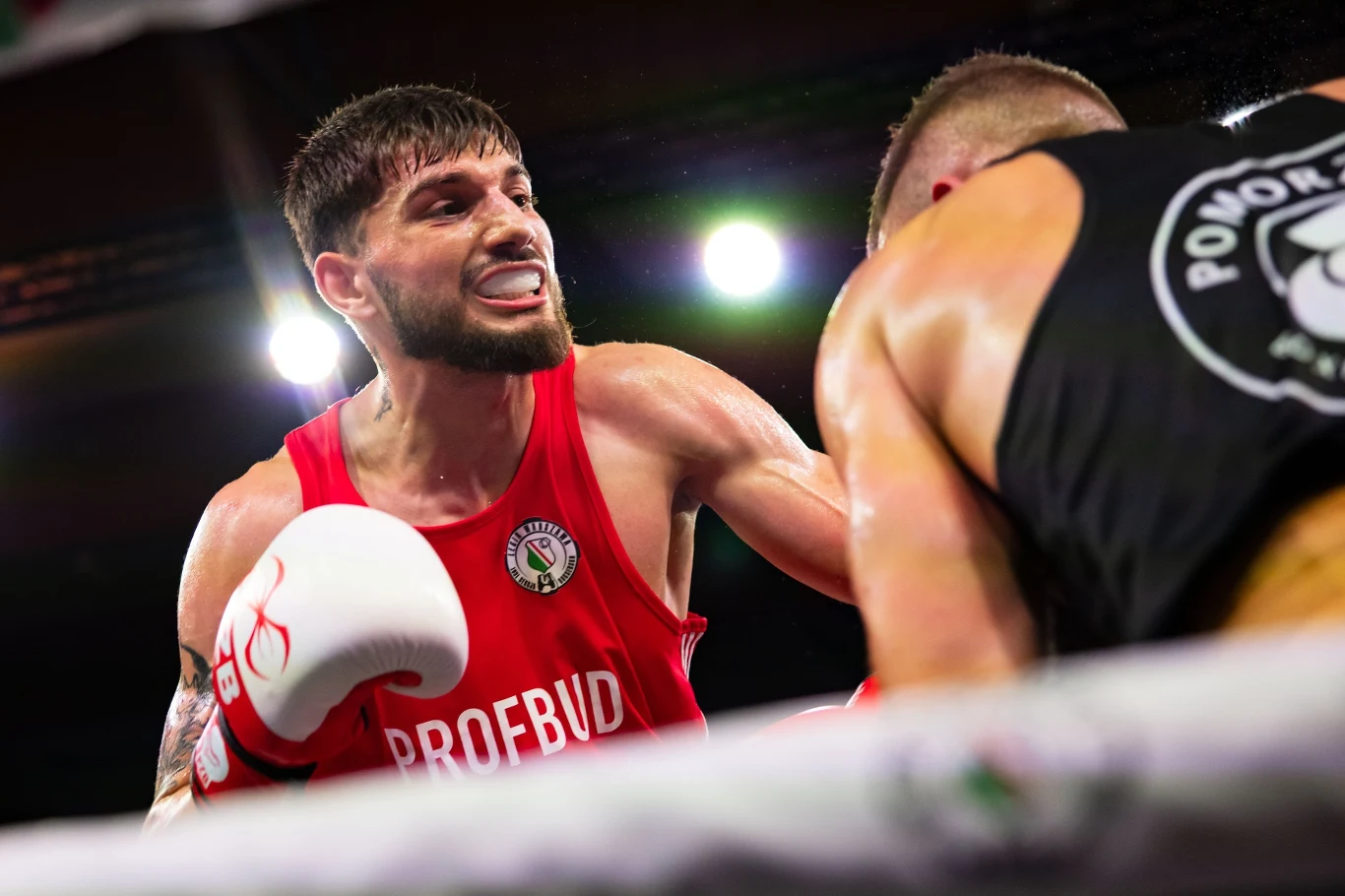Dwóch bokserów w trakcie walki na ringu, jeden z nich ubrany w czerwony strój, zaciśnięte pięści i skoncentrowane spojrzenie, drugi lekko pochylony, w czarnej koszulce z logo na plecach, w tle jasne światła areny.