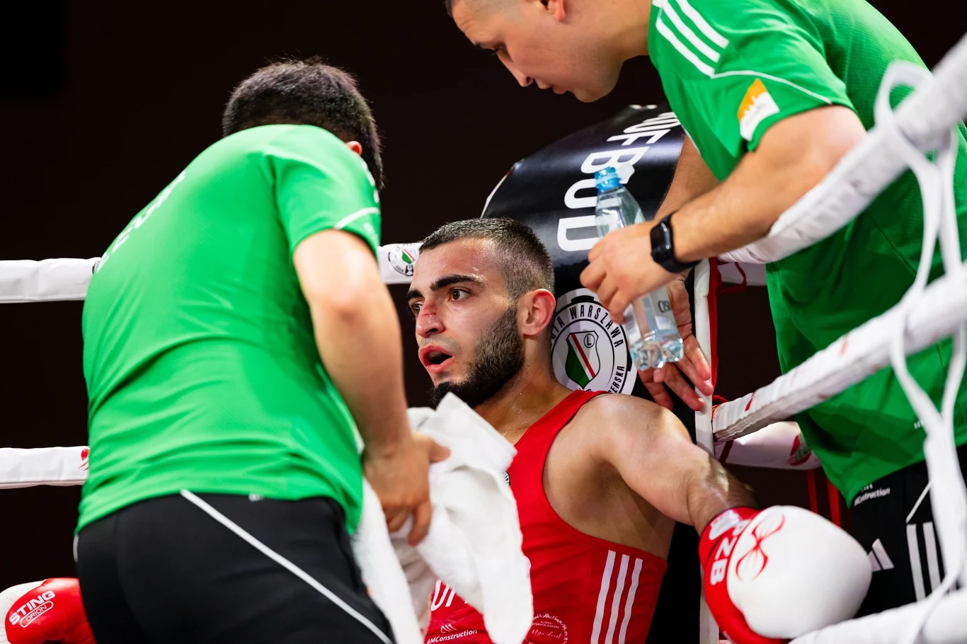 Bokser w czerwonym stroju siedzi w narożniku ringu podczas przerwy między rundami, otoczony przez dwóch trenerów w zielonych koszulkach podających mu wodę i ręcznik.