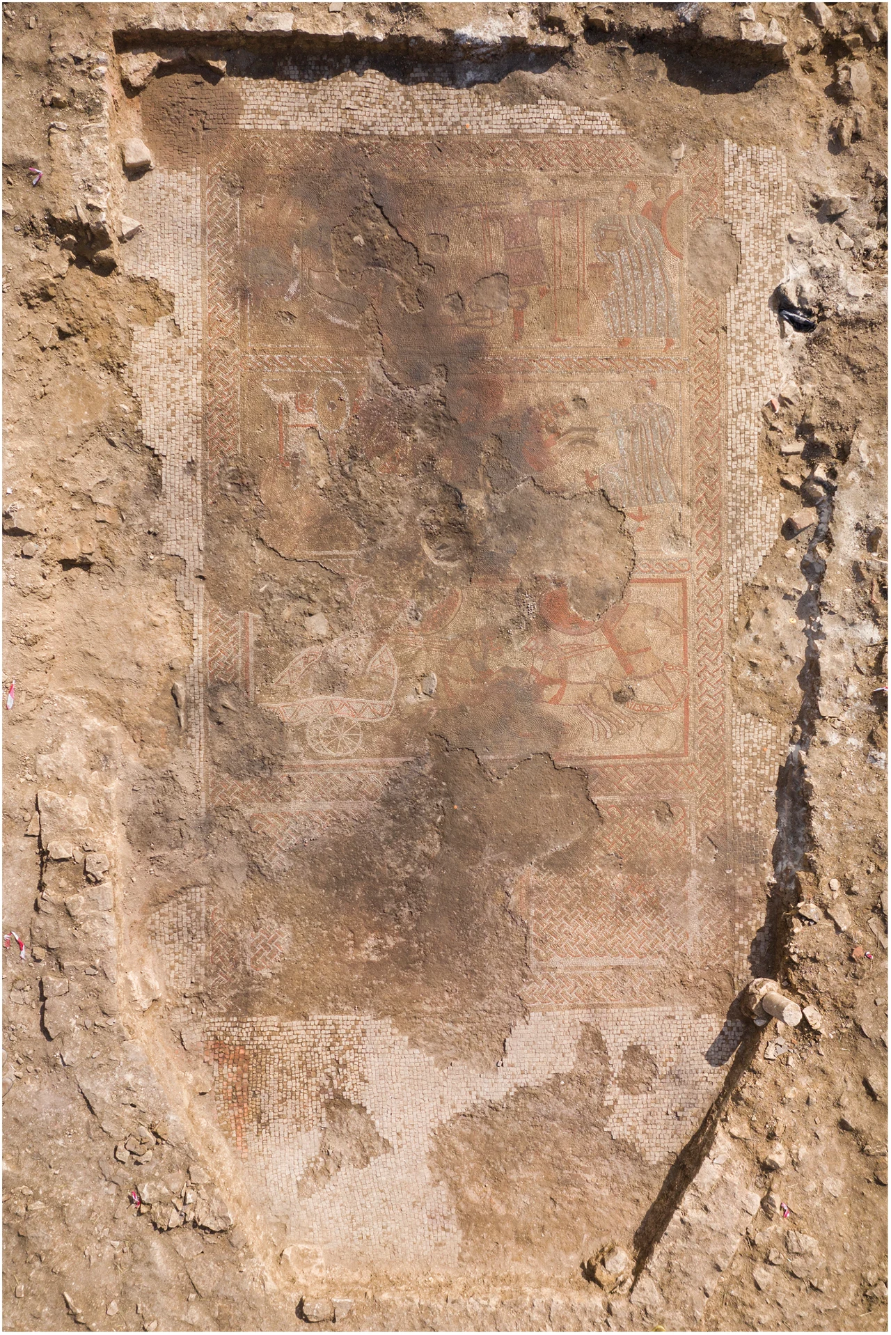 Odkryta w ziemi starożytna mozaika o prostokątnym kształcie, częściowo uszkodzona i zabrudzona ziemią, otoczona wykopem archeologicznym.