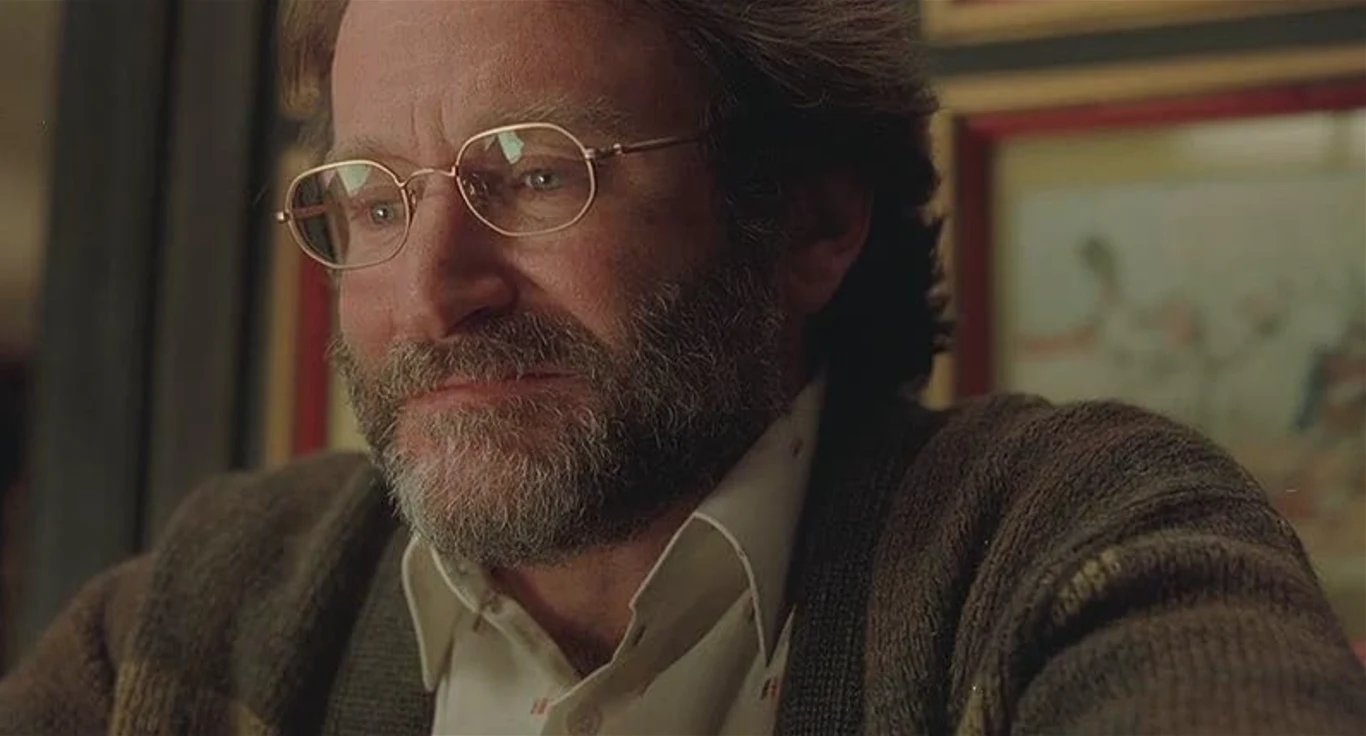 Robin Williams w filmie "Buntownik z wyboru" Mężczyzna w średnim wieku w okularach z brodą i w swetrze, wyrażający emocje smutku lub zadumy, siedzi przy stole w ciepłym, domowym wnętrzu.