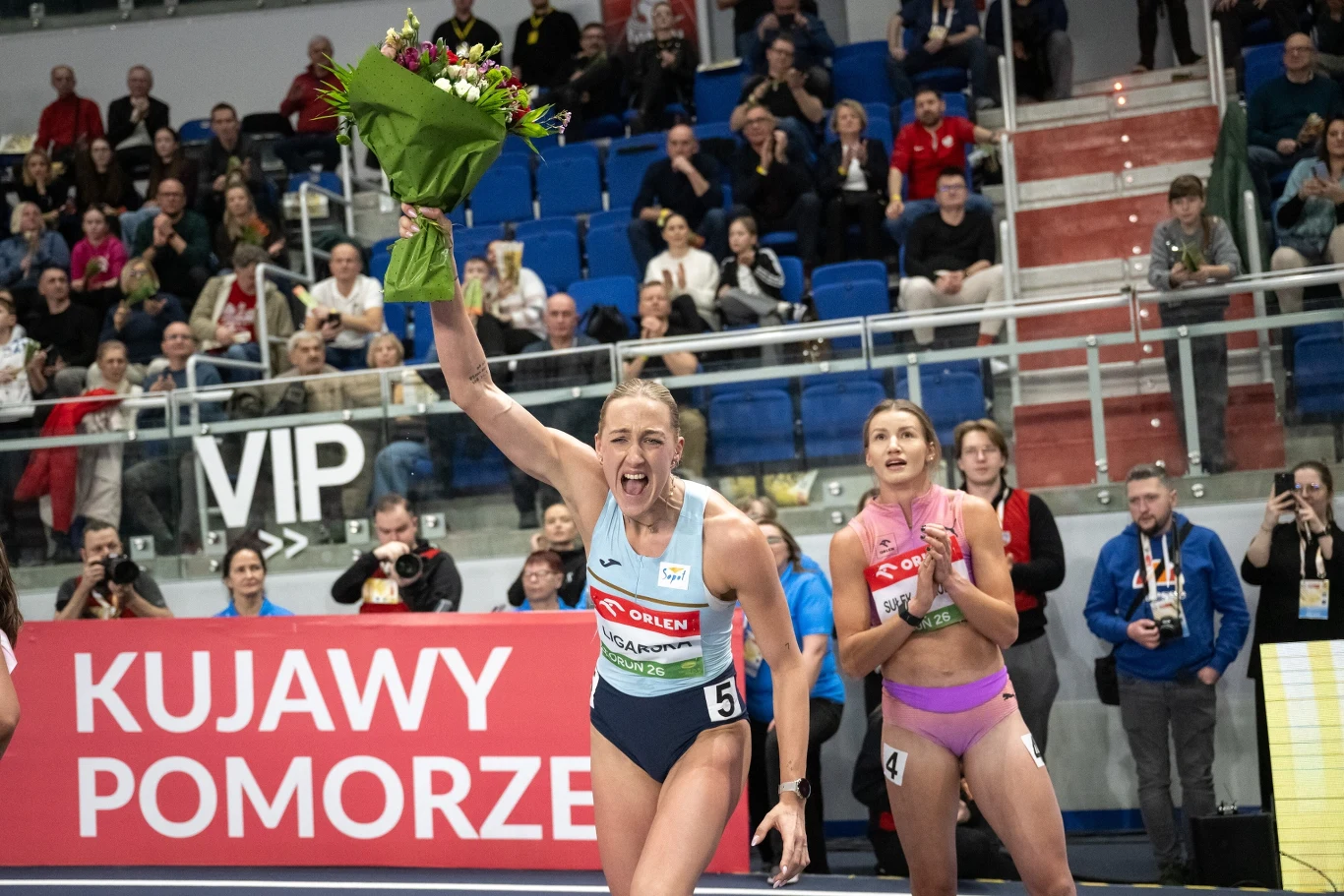 Lekkoatletka w stroju sportowym trzyma w górze bukiet kwiatów i świętuje zwycięstwo na tle pełnej trybuny kibiców w hali. W tle druga zawodniczka oraz transparent z napisem Kujawy Pomorze.