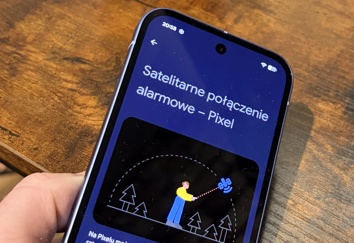 Smartfon trzymany w dłoni wyświetlający ekran z instrukcją dotyczącą satelitarnych połączeń alarmowych w telefonach Pixel, na ilustracji widoczna jest postać z telefonem w otoczeniu drzew.