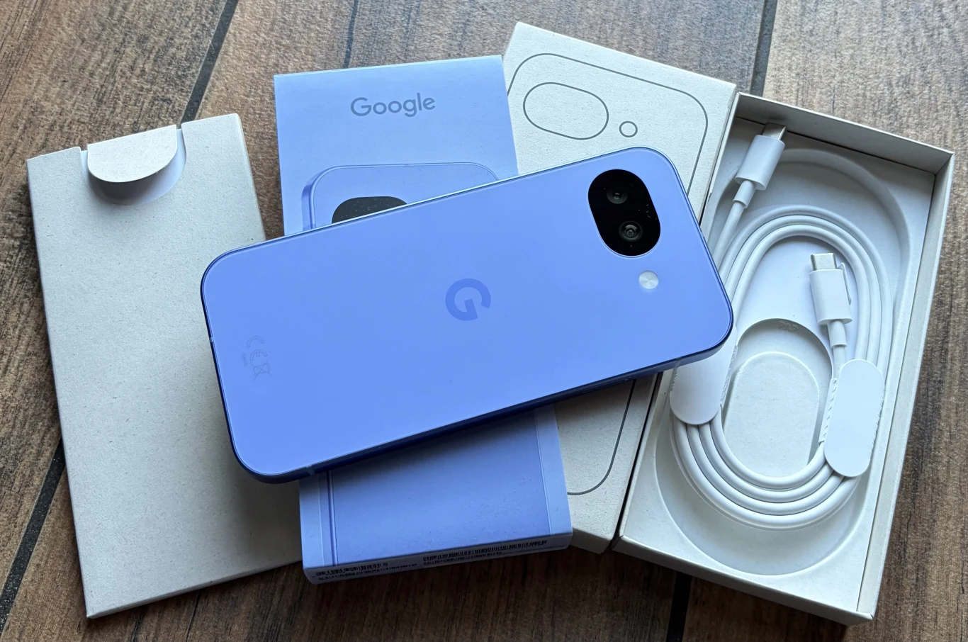 Fioletowy smartfon Google leży na swoim opakowaniu obok przewodu USB-C i papierowej dokumentacji, wszystko ułożone na drewnianej powierzchni.