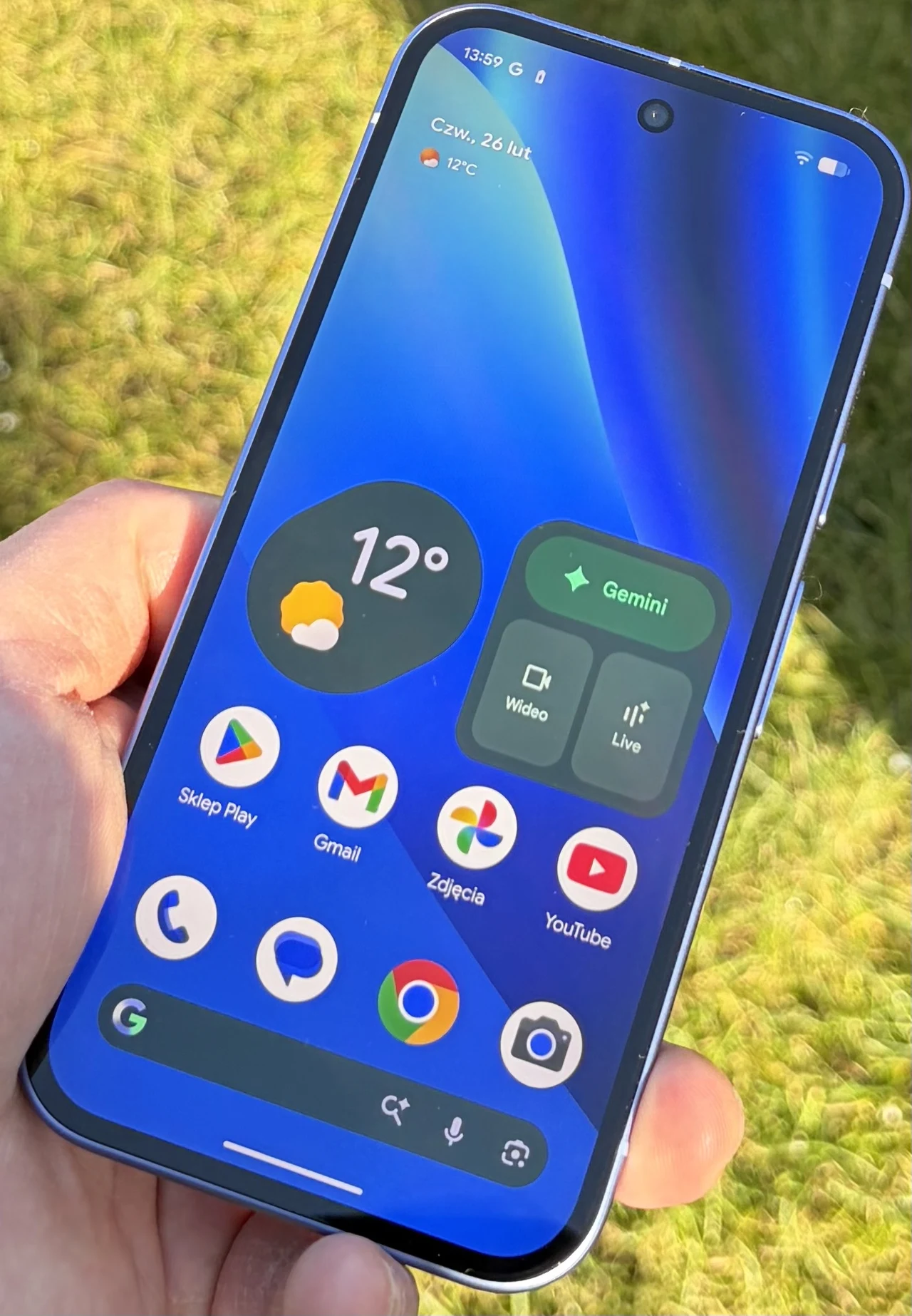 Nowoczesny smartfon trzymany w lewej dłoni na tle zielonej trawy, z włączonym ekranem prezentującym pogodę, skróty do aplikacji, pasek wyszukiwania Google oraz widoczne powiadomienia.