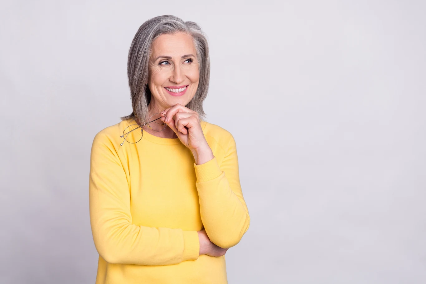 Kobieta w średnim wieku z siwymi włosami, uśmiechnięta, trzymająca okulary w jednej ręce i ubrana w żółty sweter, patrząca w bok na jasnym tle.