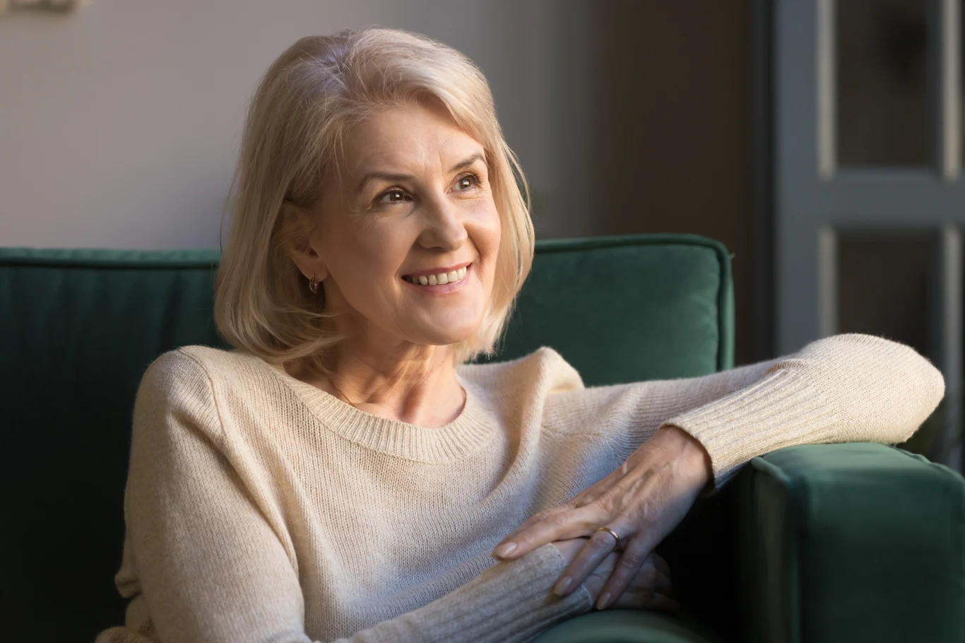 Starsza kobieta z jasnymi włosami uśmiecha się, siedząc wygodnie na zielonej sofie, ubrana w kremowy sweter, patrzy w bok z pozytywnym wyrazem twarzy.