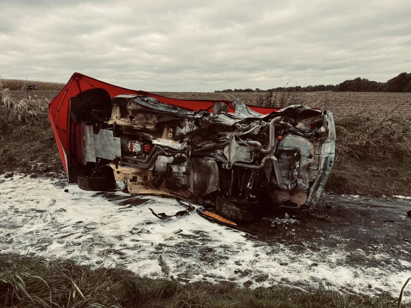 Podczas Rajdu Nyskiego doszło do wypadku, w którym Maciej Krzysik został poważnie ranny Spalony i przewrócony na bok wrak czerwonego samochodu osobowego na poboczu wiejskiej drogi, otoczony śladami piany gaśniczej oraz zniszczonym terenem, w tle pole uprawne i pochmurne niebo.