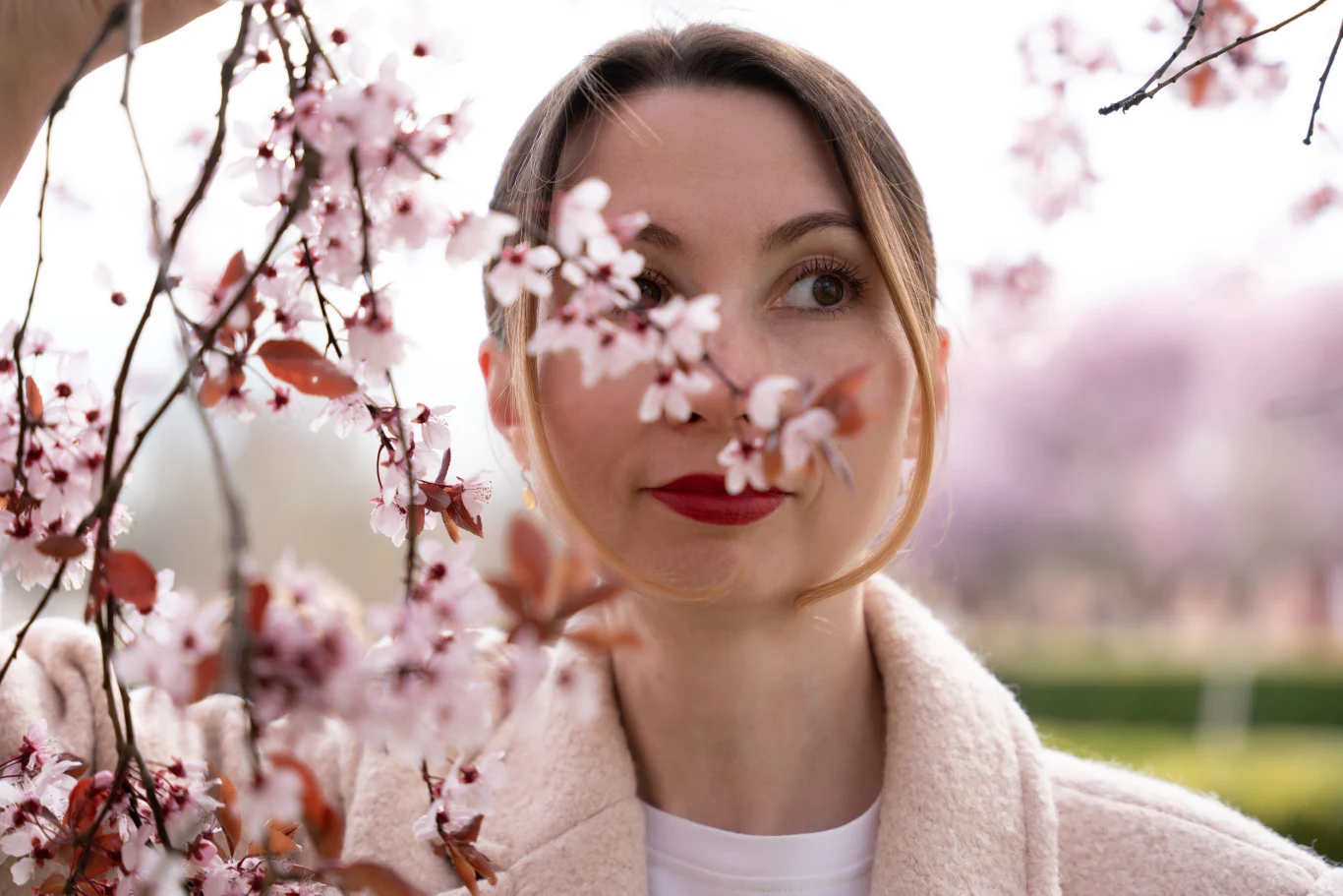 Sprawdź, co przyniesie marzec Kobieta o jasnej cerze i czerwonych ustach stoi wśród kwitnących gałęzi drzewa, delikatne różowe kwiaty są blisko jej twarzy, otaczają ją naturalną ramą.