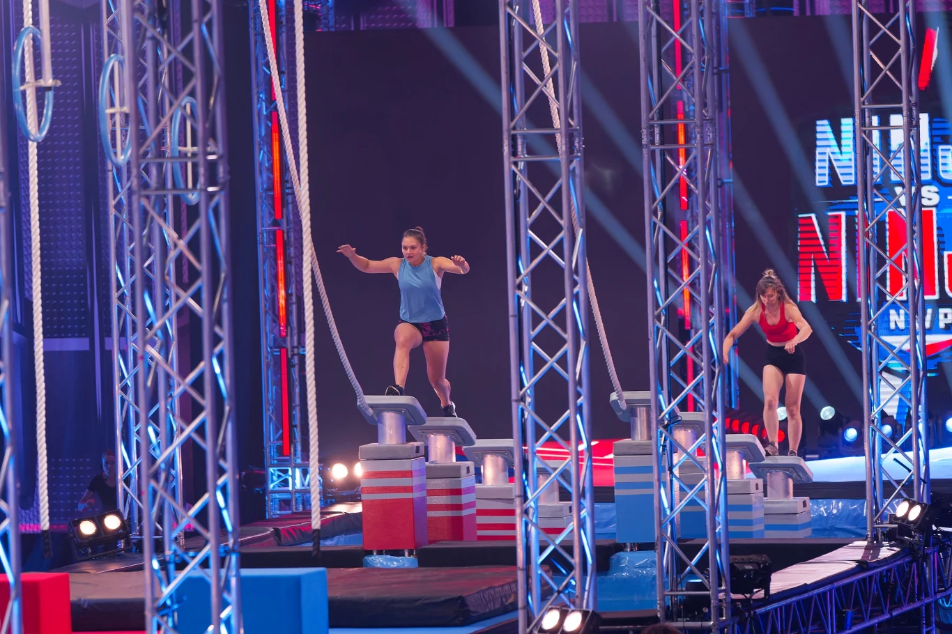 Dwie kobiety pokonują tor przeszkód w stylu programu Ninja Warrior, skacząc po platformach i chwytając liny, otoczone kolorowymi światłami oraz metalowymi konstrukcjami.