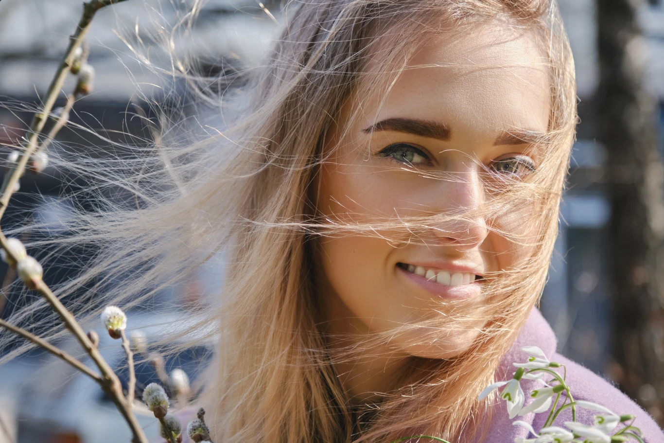 Marzec pod znakiem gwiazd. Co czeka twój znak zodiaku w nadchodzącym miesiącu? Uśmiechnięta młoda kobieta z jasnymi włosami powiewającymi na wietrze, stojąca w pobliżu gałązki z pąkami wierzbki.