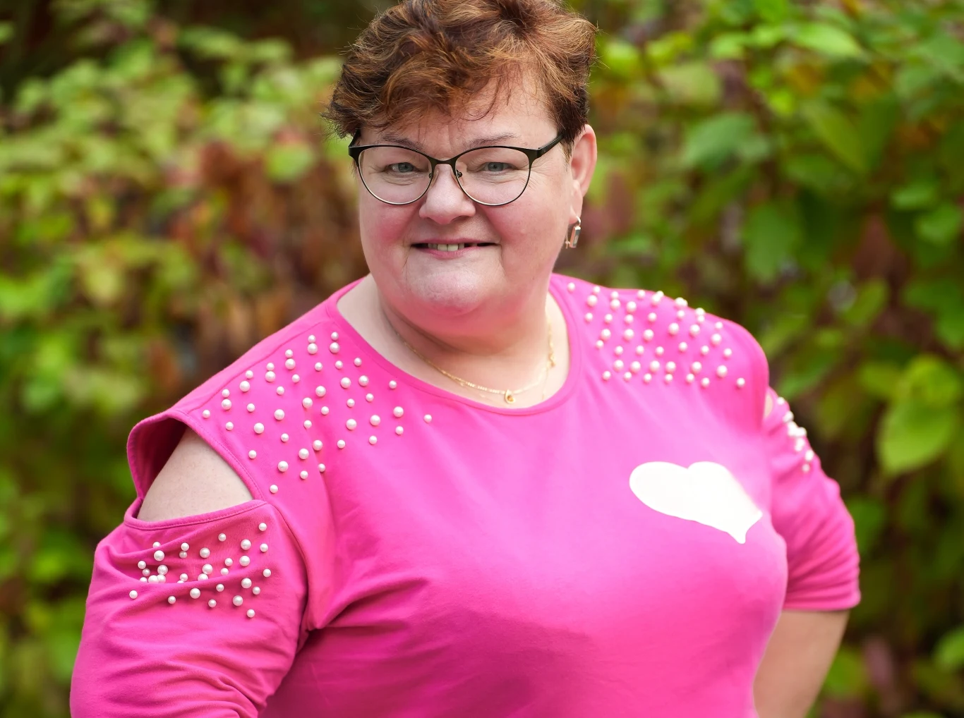 Lila z "Sanatorium miłości" Kobieta w średnim wieku stojąca na tle zielonych roślin, uśmiechnięta, ubrana w różową bluzkę z ozdobnymi perełkami i białym sercem. Ma krótkie, brązowe włosy i okulary.