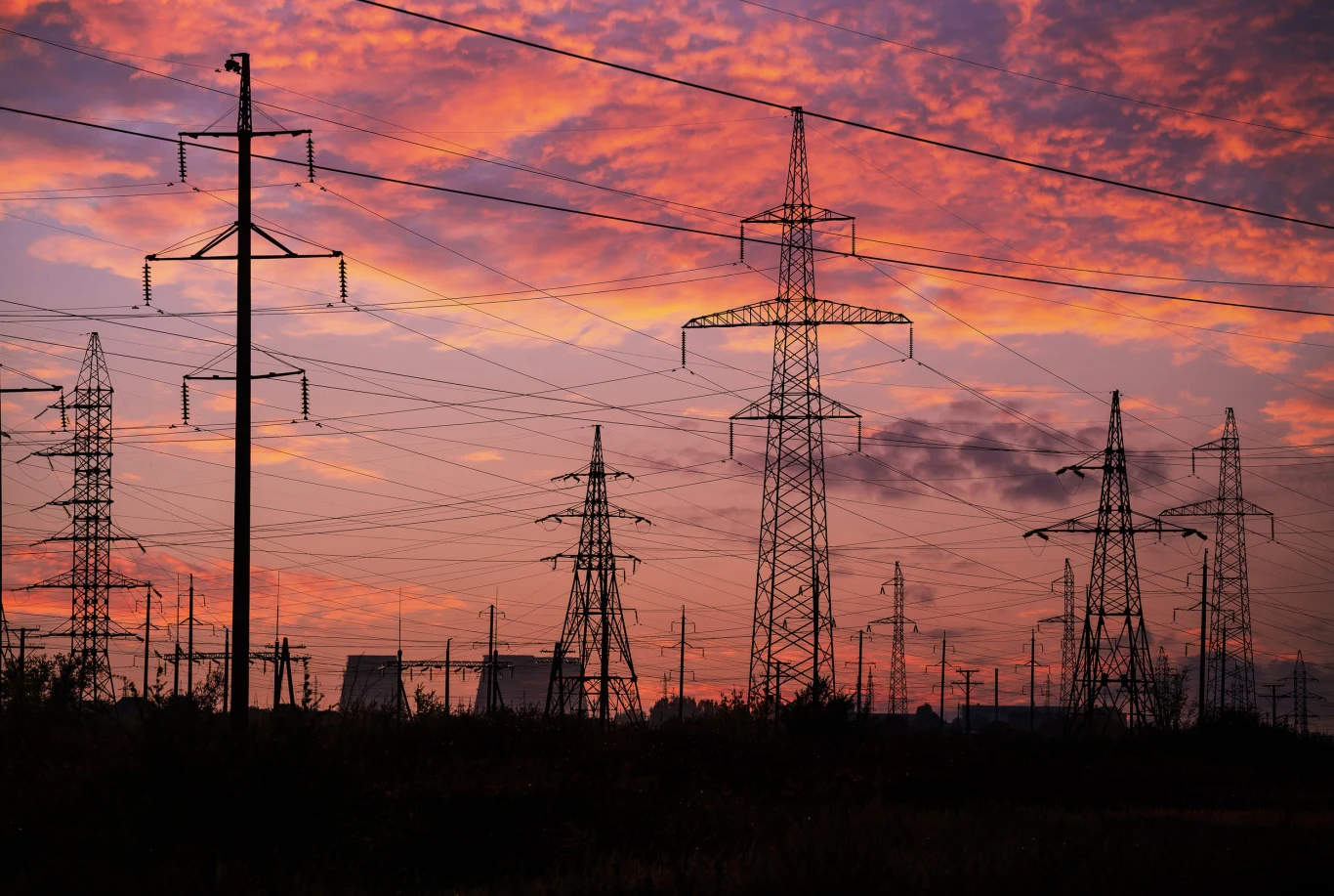 Służby w Skandynawii postawione w stan gotowości. Poważne zagrożenie ataku na sieć energetyczną. Zdj. ilustracyjne Linie wysokiego napięcia oraz słupy energetyczne na tle kolorowego, zachmurzonego nieba o zachodzie słońca, z widocznymi zabudowaniami przemysłowymi na horyzoncie.