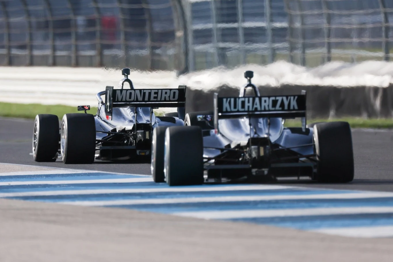 W tym sezonie Tymek Kucharczyk będzie reprezentował barwy zespołu HMD Motorsports i walczył o czołowe miejsca w serii IndyNXT Dwa bolidy wyścigowe jadą po torze asfaltowym, jadą jeden za drugim, widoczne nazwiska kierowców na tylnej części pojazdów, metalowe barierki ochronne w tle.