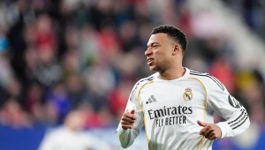 Piłkarz Realu Madryt w białym stroju z widocznym logo klubu i sponsora podczas meczu na boisku, rozmyte tło stadionu z kibicami.