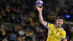 Zawodnik ubrany w żółto-niebieski strój sportowy unosi rękę z piłką do rzutu podczas meczu piłki ręcznej, na drugim planie widoczna jest publiczność na trybunach.