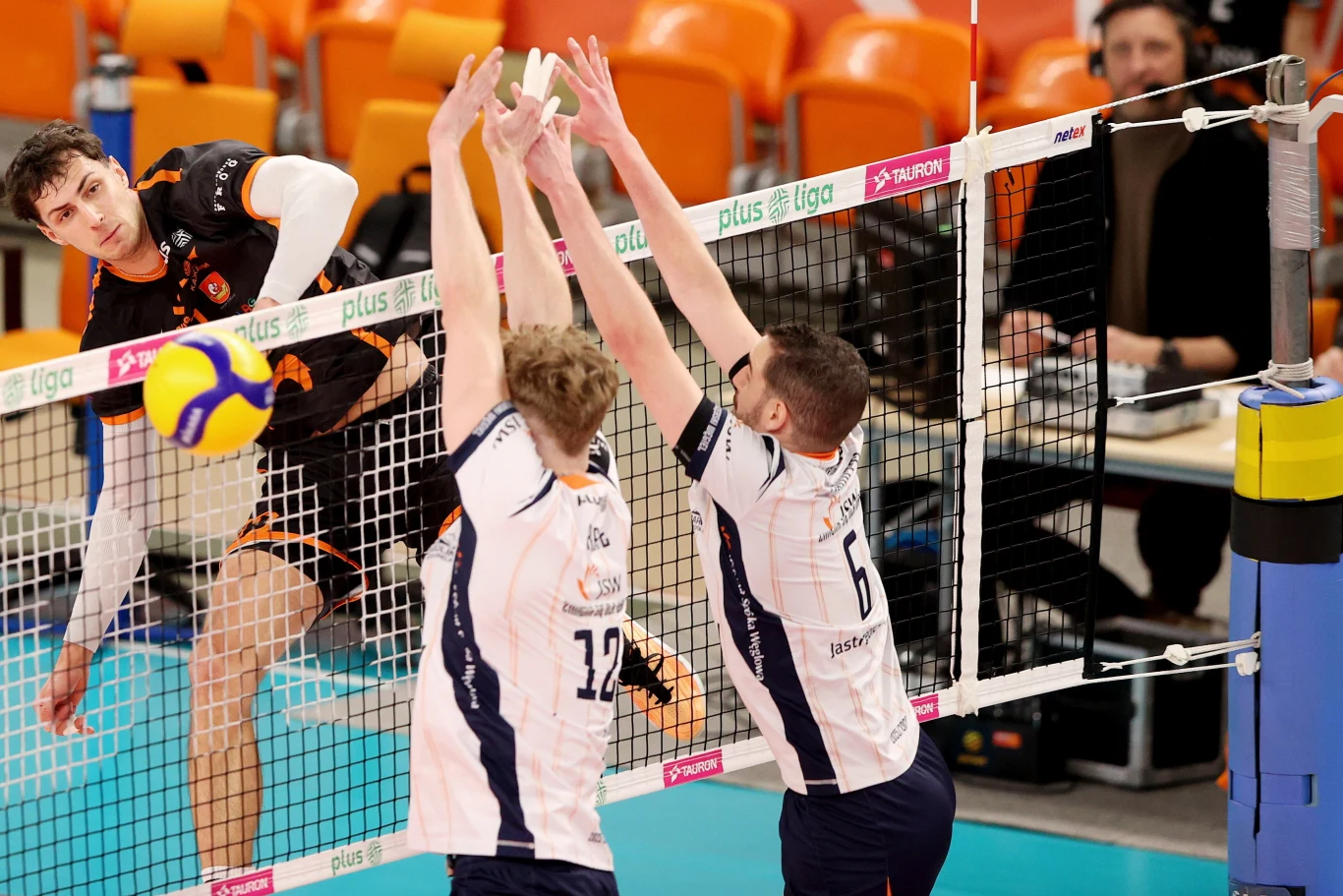 Julius Firkal atakuje w meczu JSW Jastrzębski Węgiel - Barkom Każany Lwów Zawodnik w czarno-pomarańczowym stroju wykonuje atak siatkarski, podczas gdy dwóch przeciwników w białych koszulkach próbuje go zablokować przy siatce. W tle widać pomarańczowe krzesełka i sędziego stolikowego.