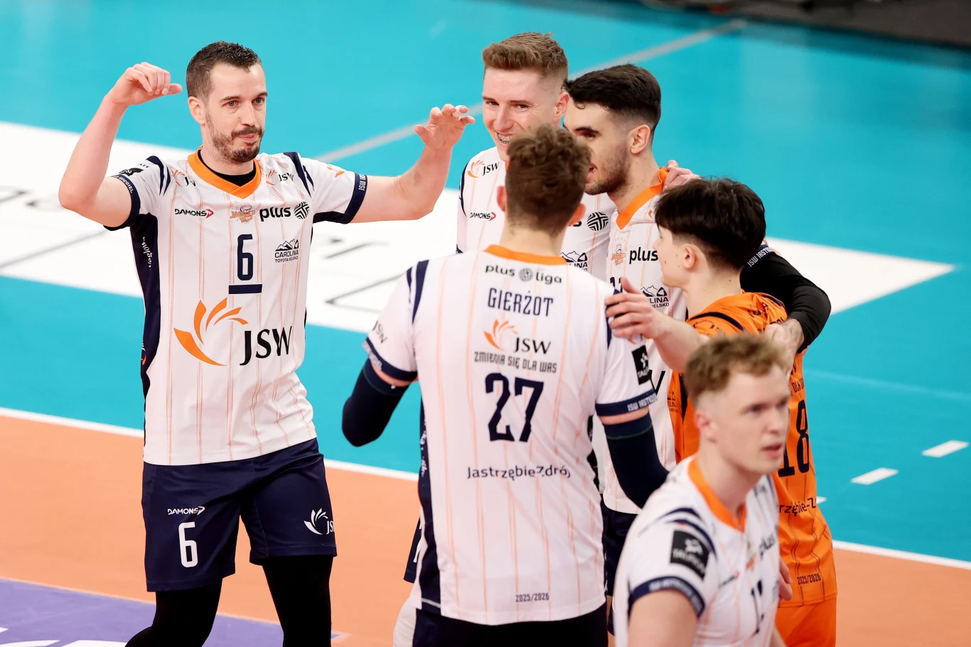 Siatkarze JSW Jastrzębskiego Węgla Grupa siatkarzy w białych i pomarańczowych strojach drużyny JSW świętuje punkt na boisku sportowym, otoczona niebieskim parkietem.